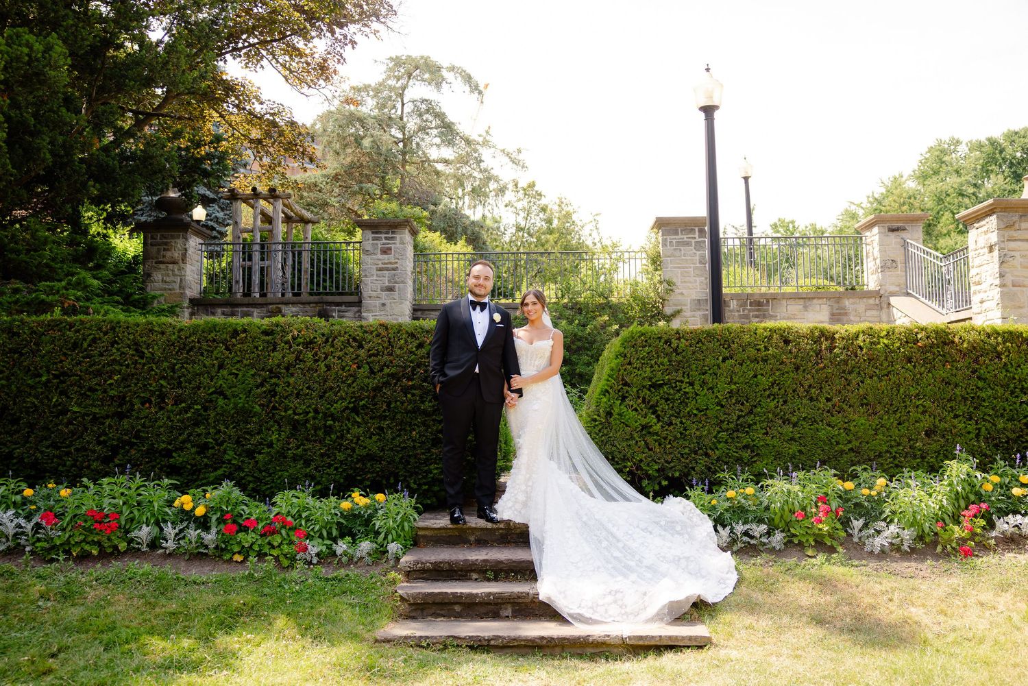 A romantic wedding couple walks through a lush garden with blooming flowers and tall greenery on a sunny day.
