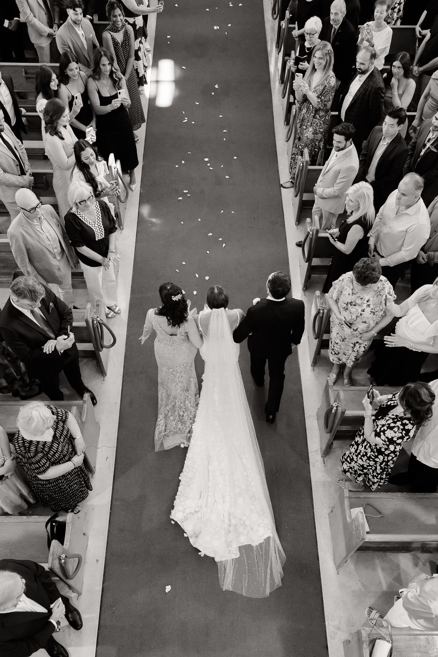 A black and white overhead view of a wedding procession walking down an aisle with guests lined up on both sides.
