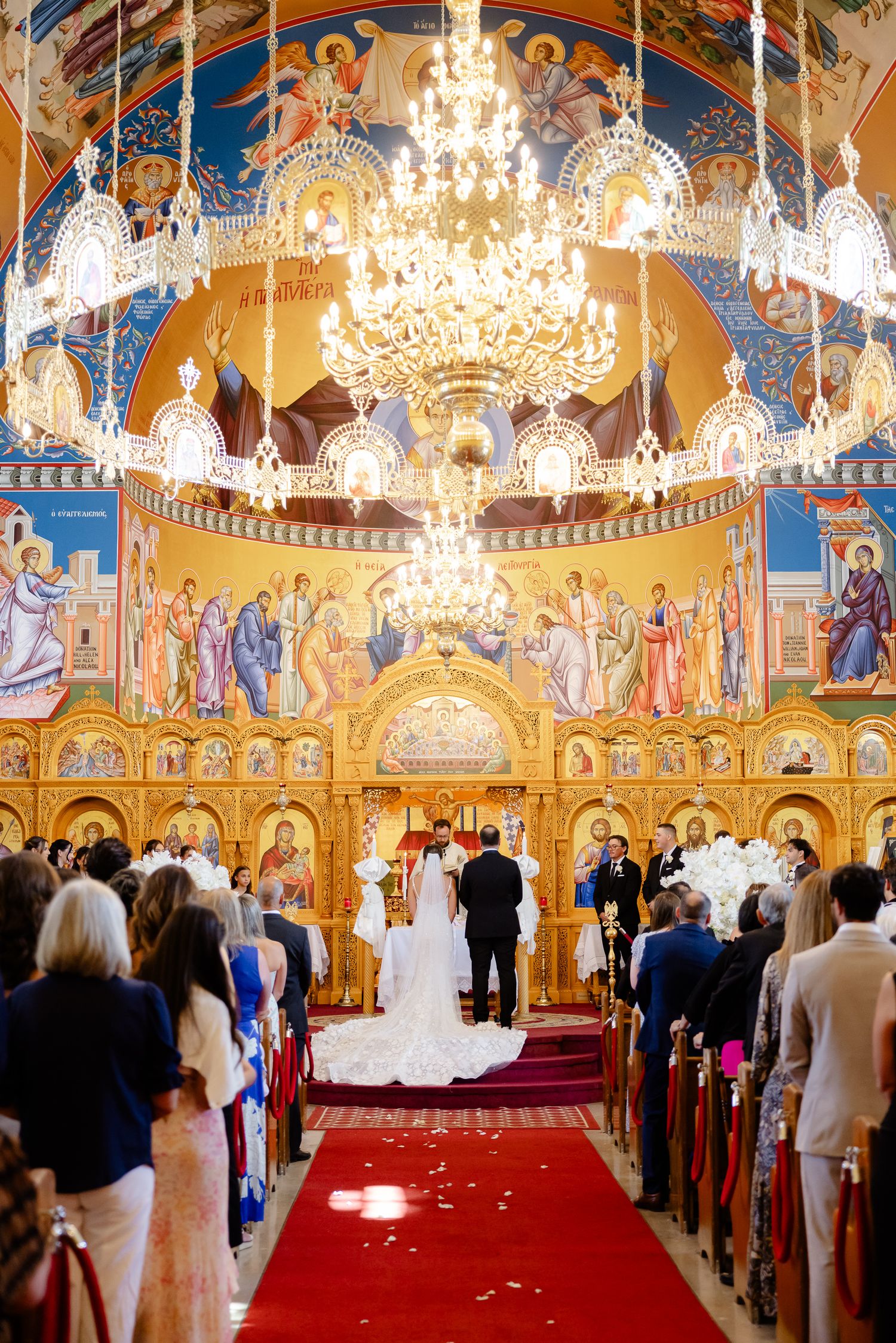 Interior of an ornate Orthodox church with golden walls, crystal chandeliers, and guests seated for a wedding ceremony.