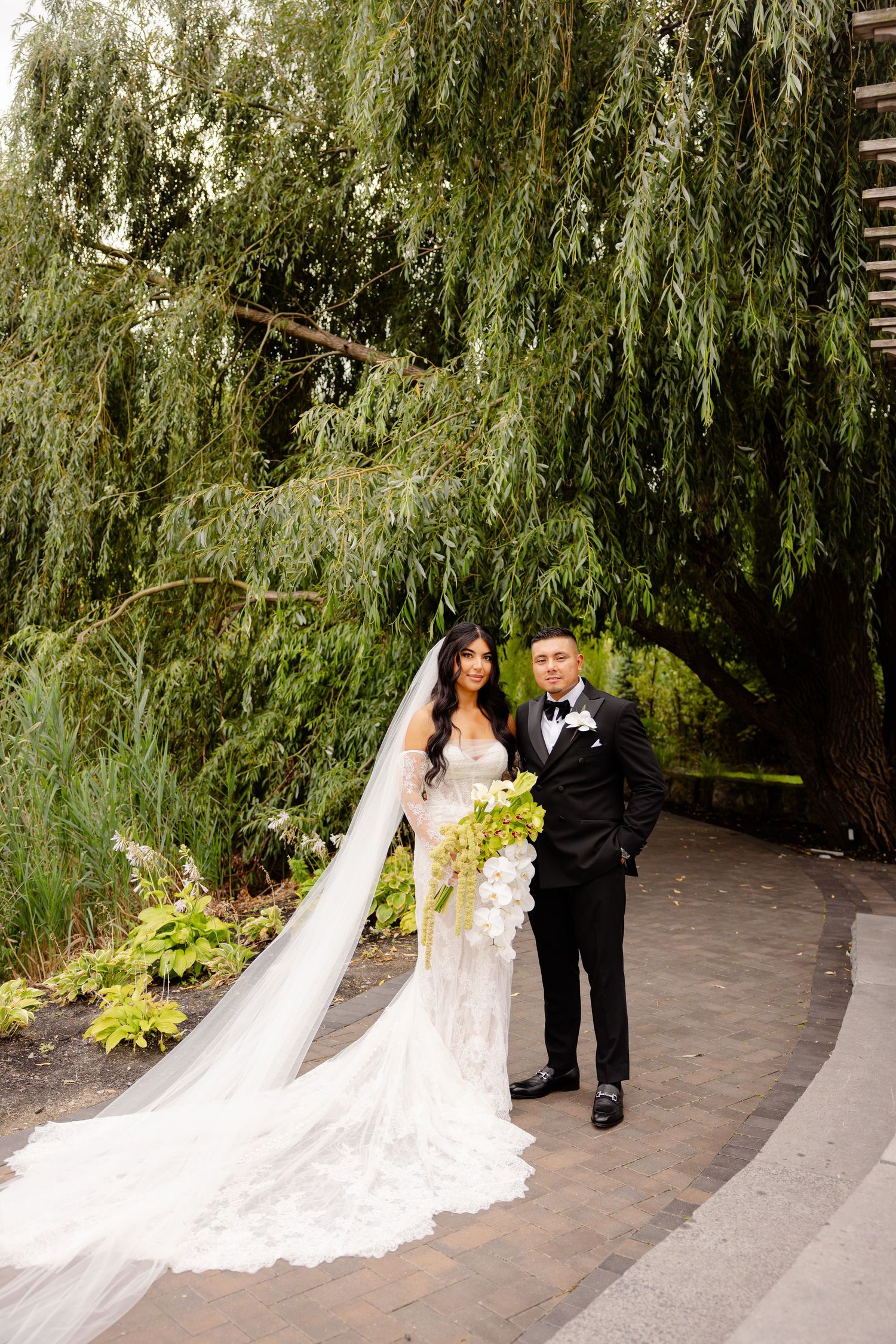 Wedding couple poses together under a romantic weeping willow tree in an outdoor garden setting.