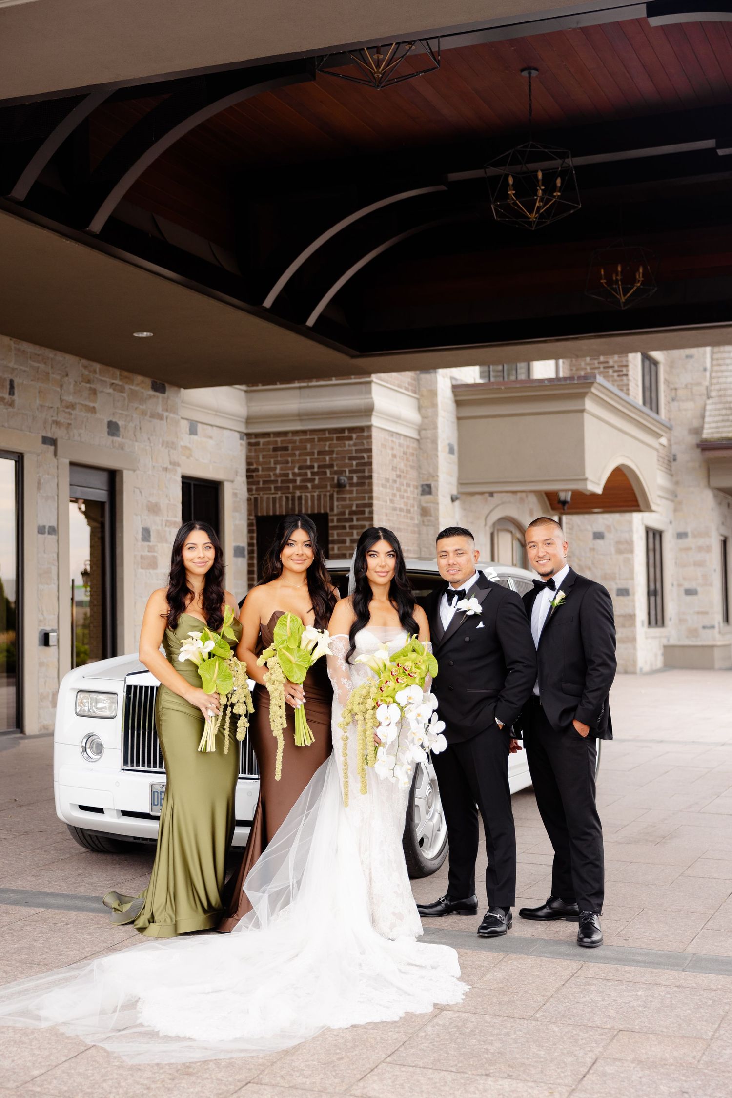 Wedding party members in light green bridesmaid dresses and formal black tuxedos pose together in a covered walkway.