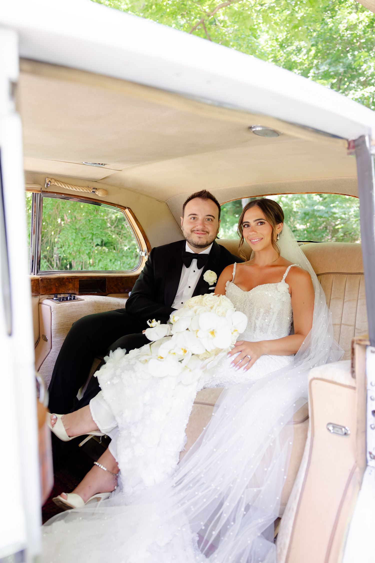 A couple in formal wedding attire sit in the back of a vintage car during their wedding celebration.