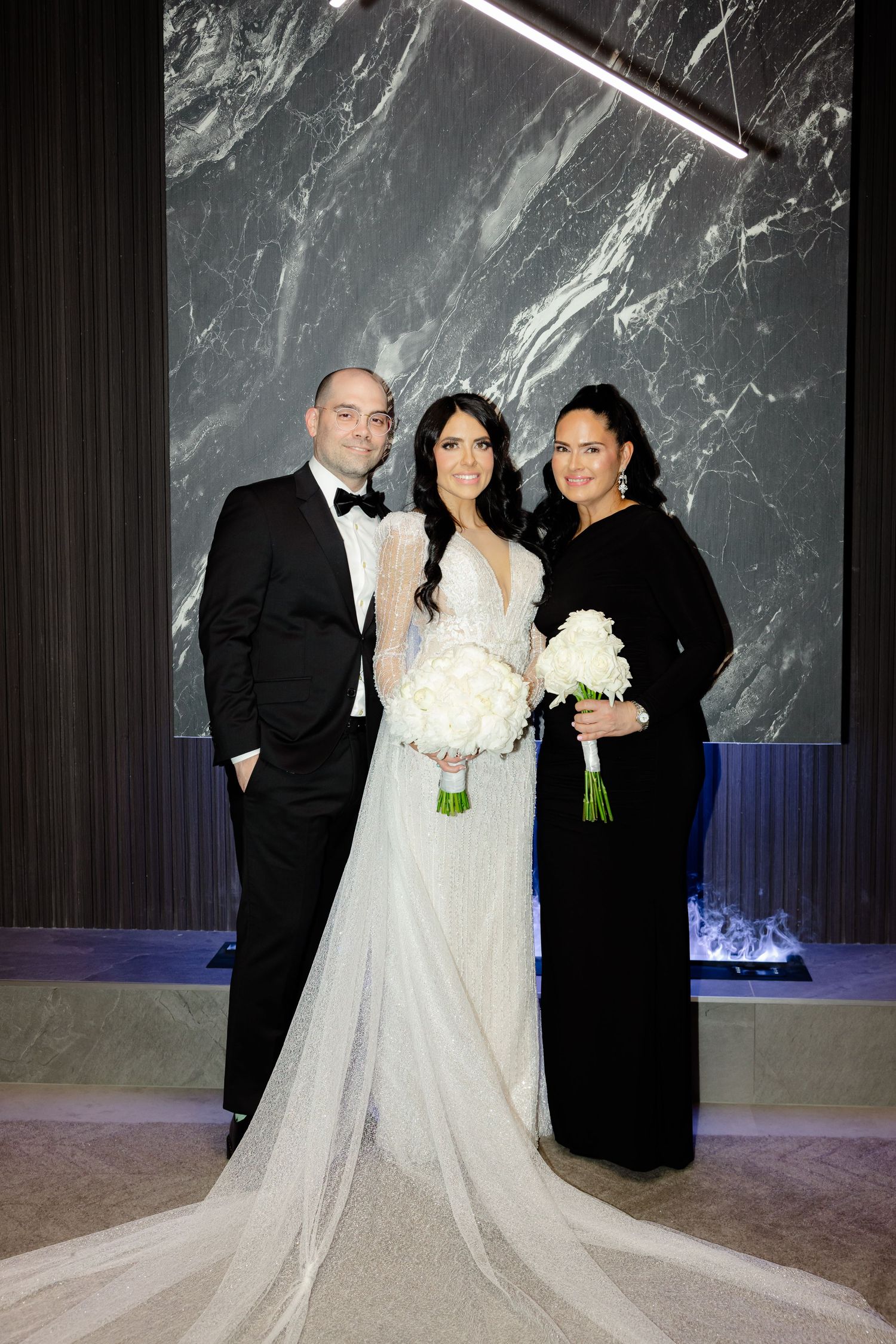 Formal wedding portrait featuring bride in flowing white gown with two guests in black attire.
