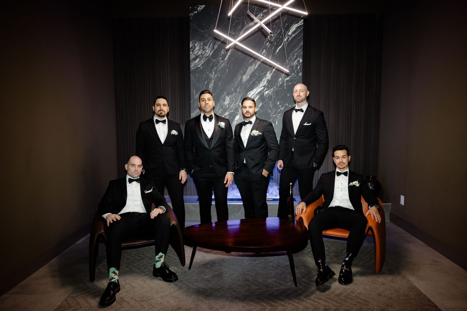 Groomsmen in formal attire seated and standing against dark wall with geometric light fixture.