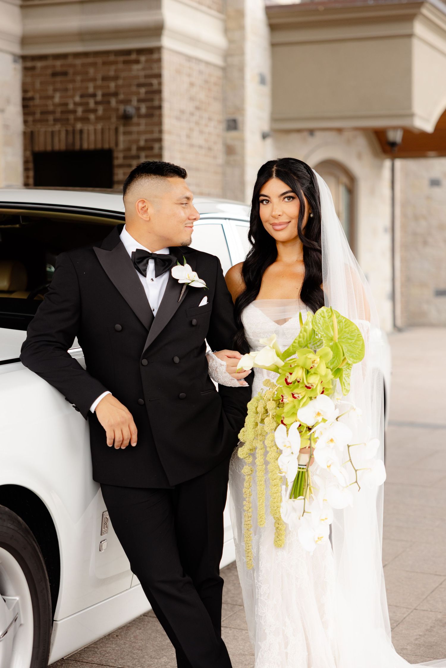 Wedding couple poses next to white luxury car with yellow bouquet and formal attire.