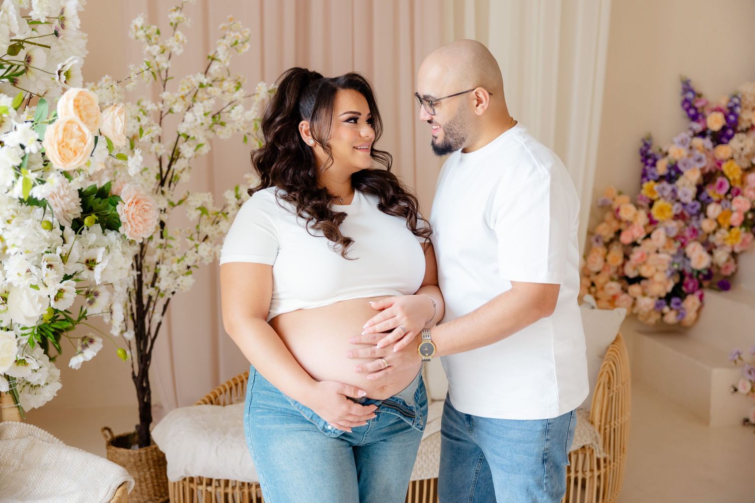 Expectant couple in white shirts sharing tender moment during maternity photoshoot with floral backdrop.