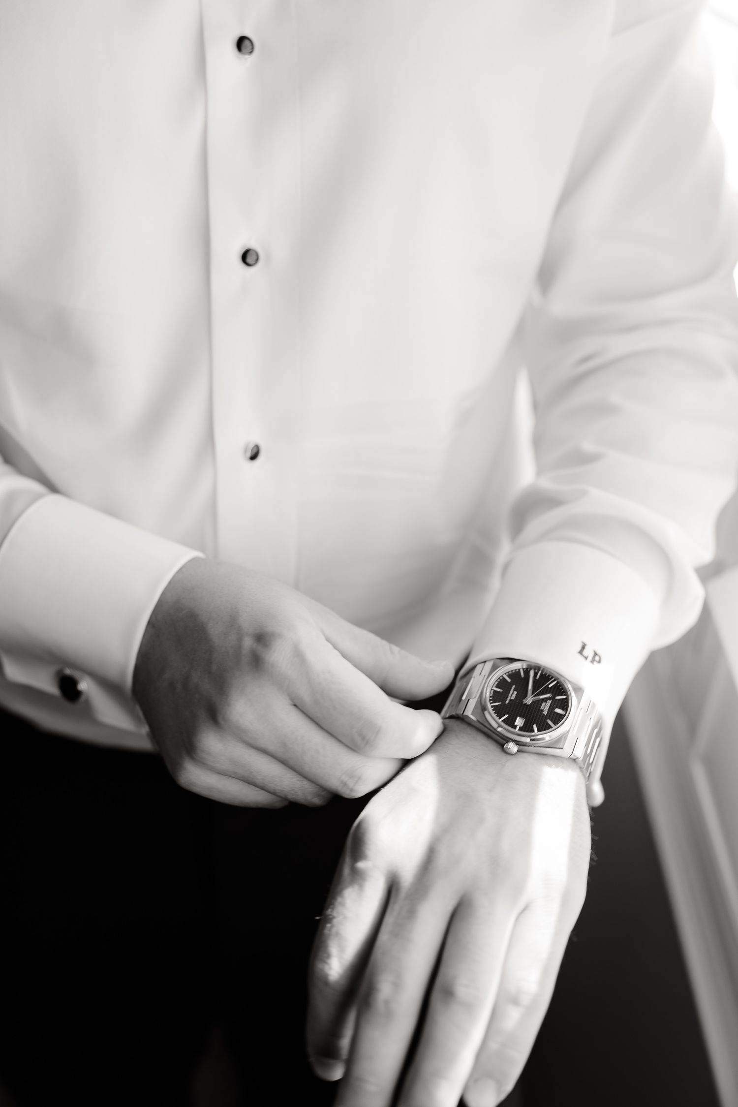Black and white photo of hands buttoning a crisp white dress shirt sleeve and adjusting a wristwatch.