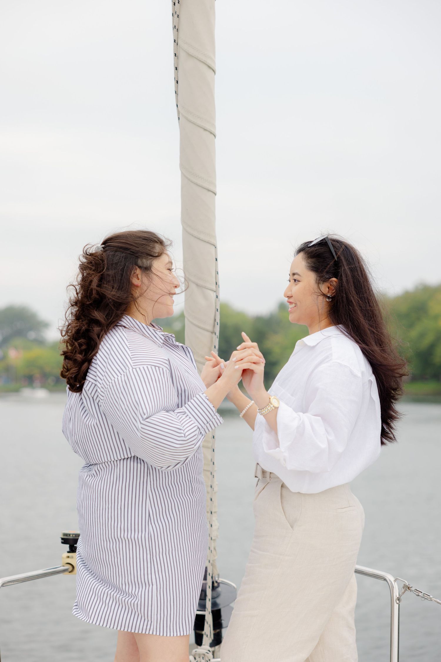Two women hold hands and smile at each other while standing on a boat deck with water and trees in the background.