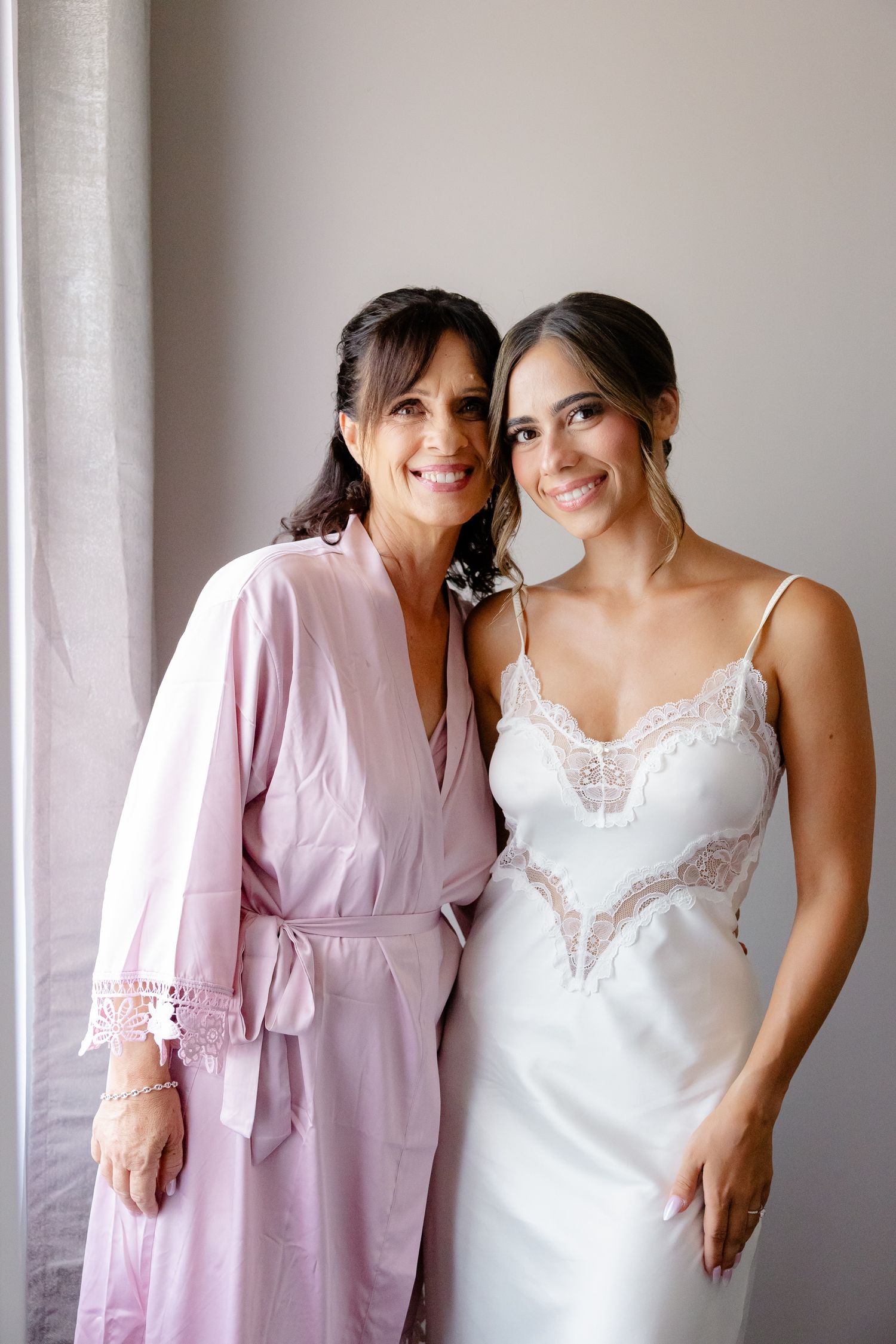 Two women pose together on a special day, one wearing a pink robe and the other in a white lace wedding dress.
