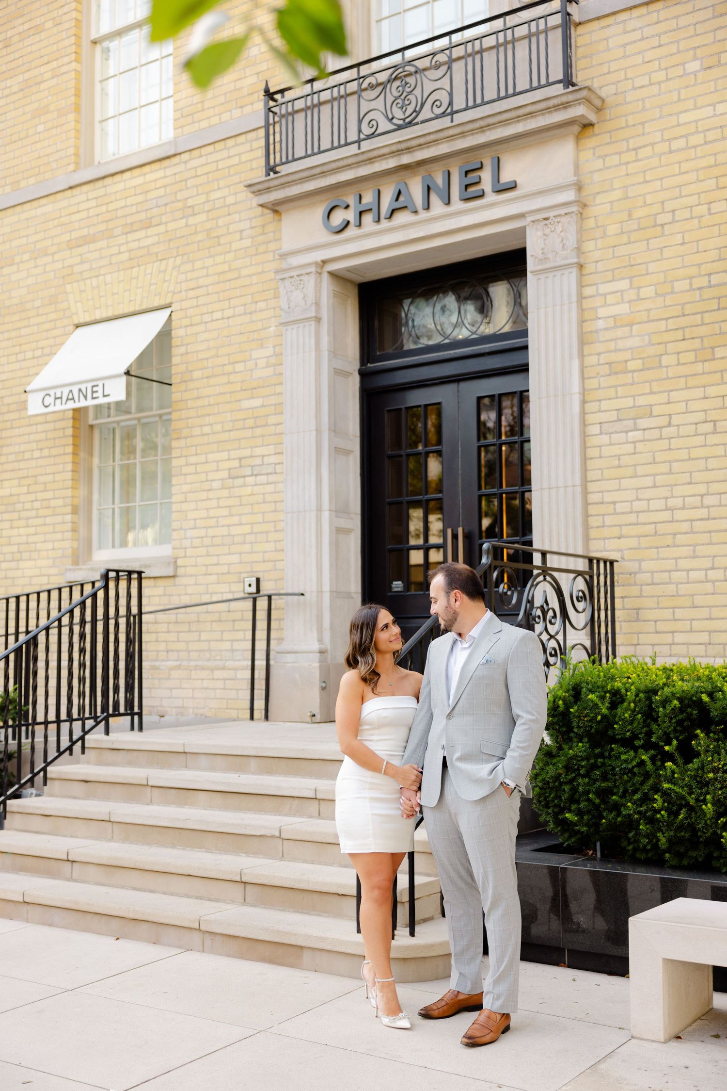 Couple in elegant attire standing on steps outside iconic Chanel storefront with cream brick facade.