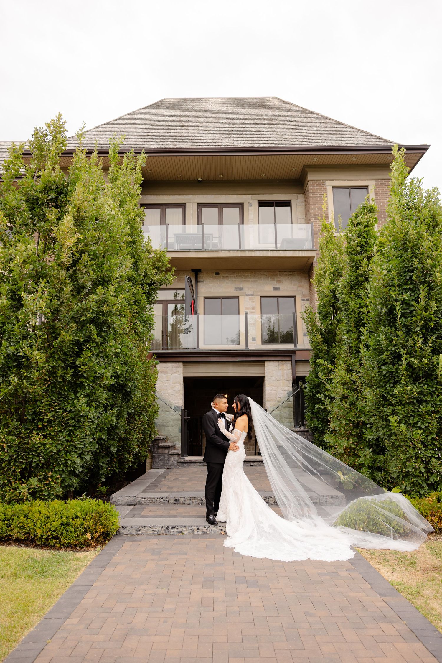 A romantic couple poses in front of a modern house with brick driveway and greenery in an elegant wedding scene.