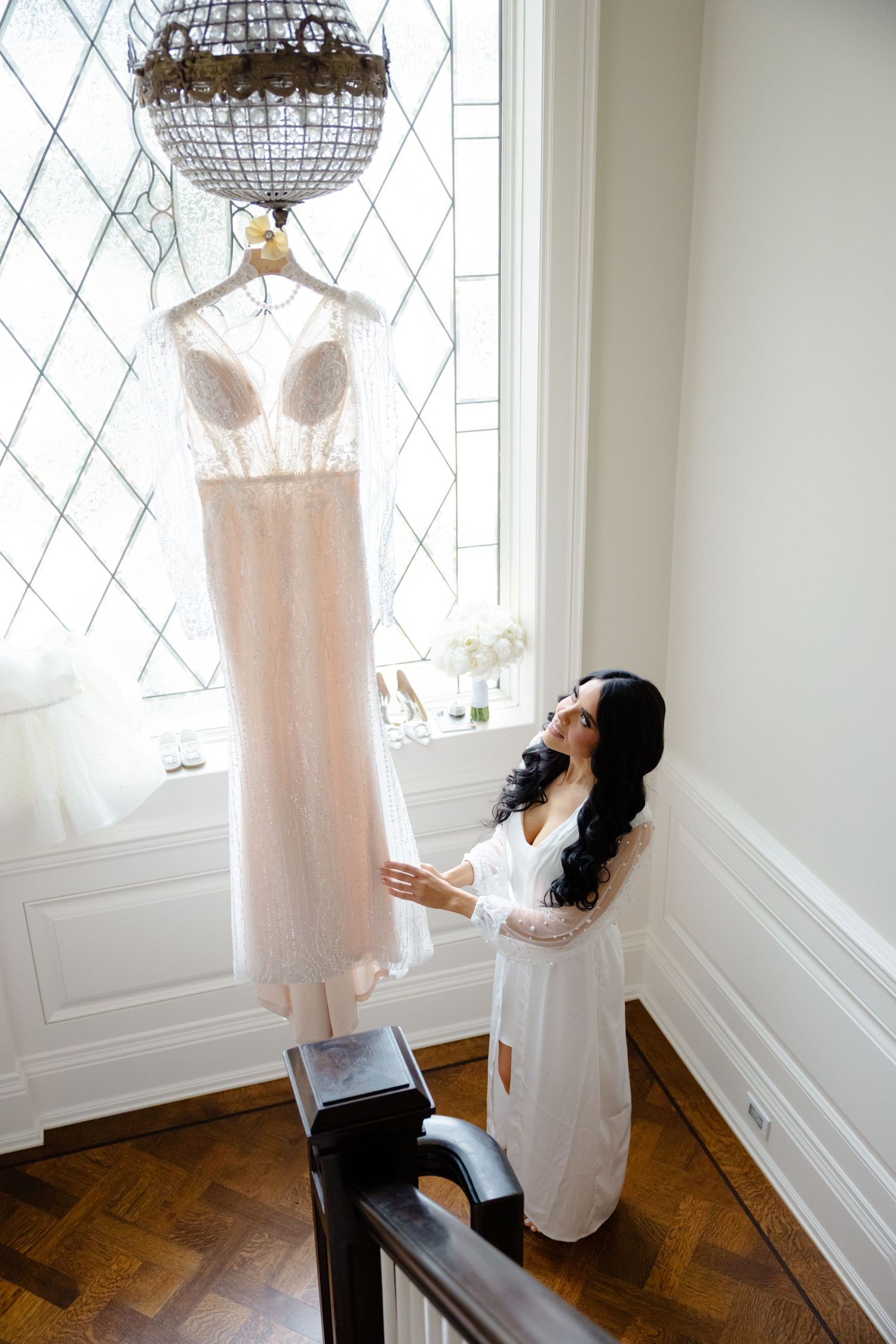 A blush pink wedding dress hangs from a crystal chandelier as someone in white reaches up towards it.