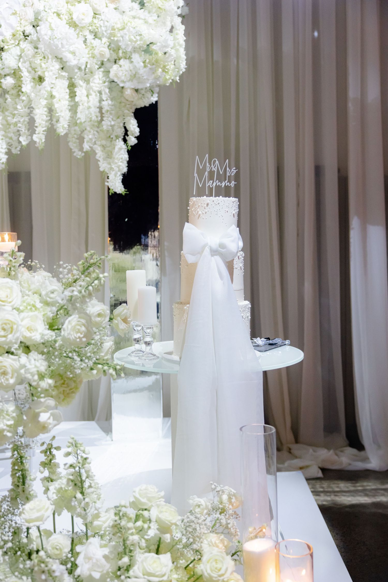 White tiered wedding cake with crystal accents and flowing bow displayed among floral arrangements and candlelight.
