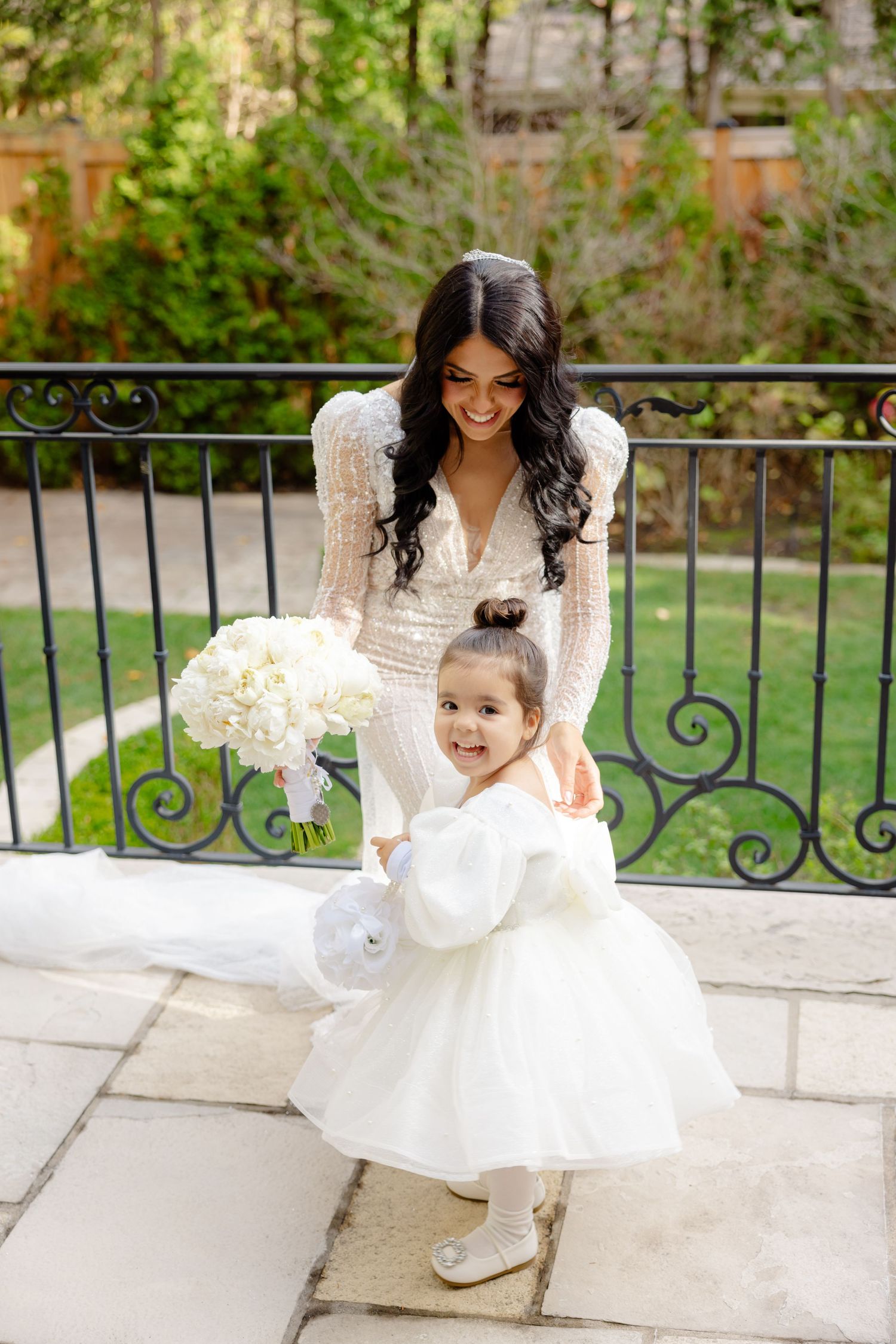 A bride in a lace gown shares a sweet moment with a small flower girl on a stone patio with ornate railings.