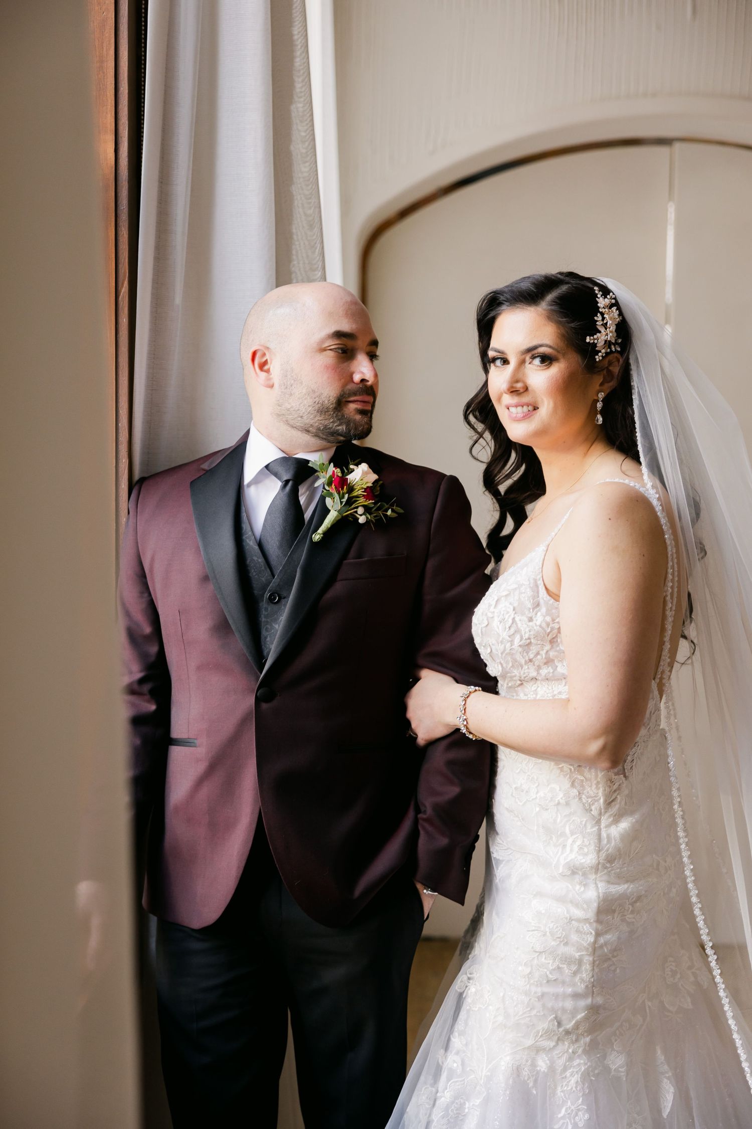 A wedding couple poses together against a light wall in formal burgundy and white attire.