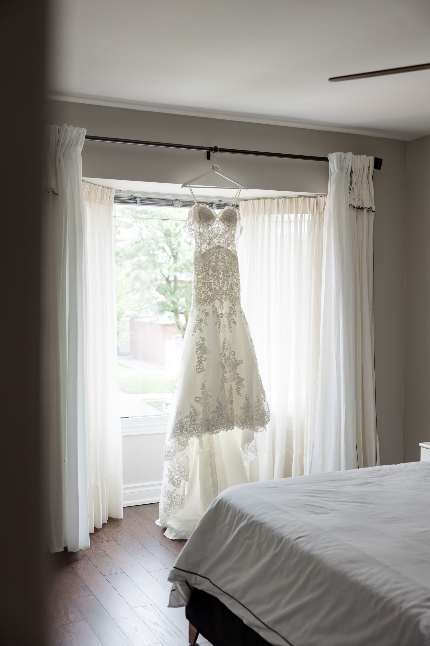 A lace white wedding dress hangs in front of a sunlit window with white curtains.