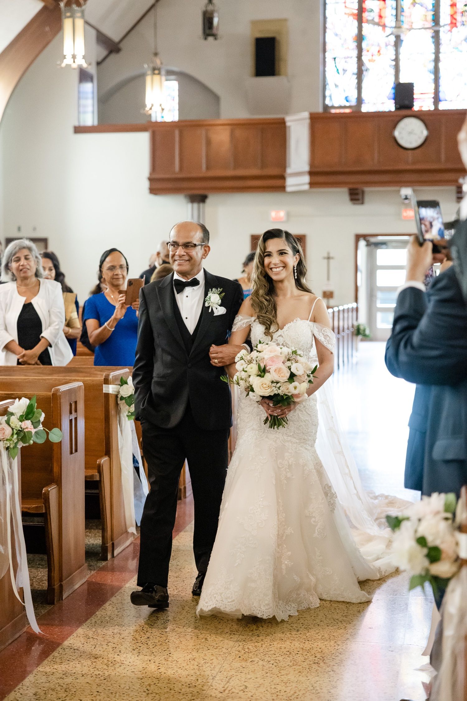 A wedding processional in a traditional church with wooden pews decorated with floral arrangements.