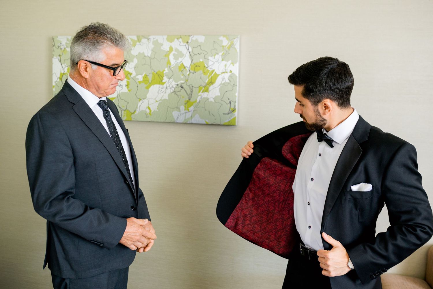 Two men in formal suits examining the lining of a tuxedo jacket during wedding preparations.