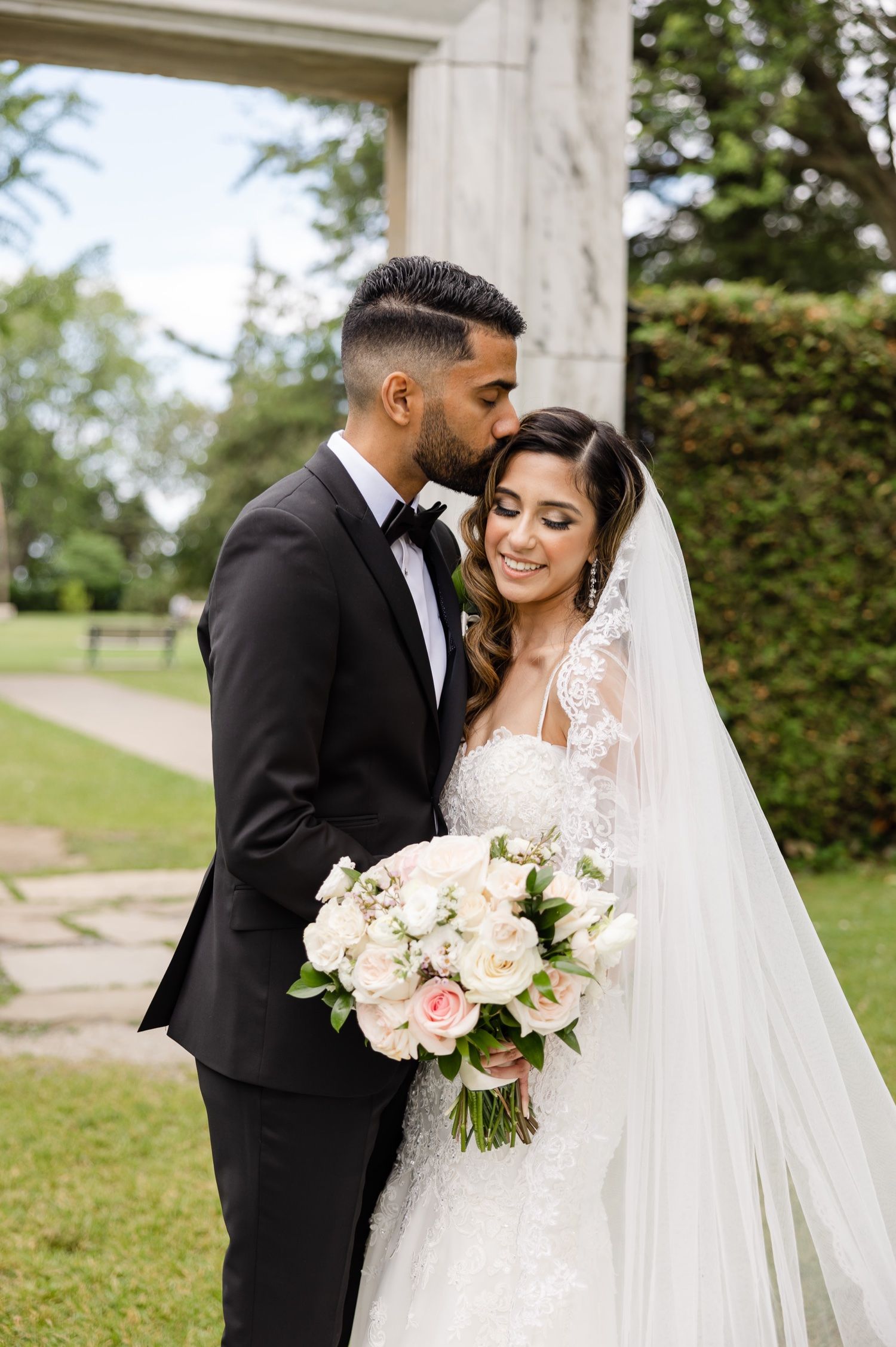 A romantic wedding portrait of a couple with the bride holding a soft pink and white rose bouquet in a garden setting.