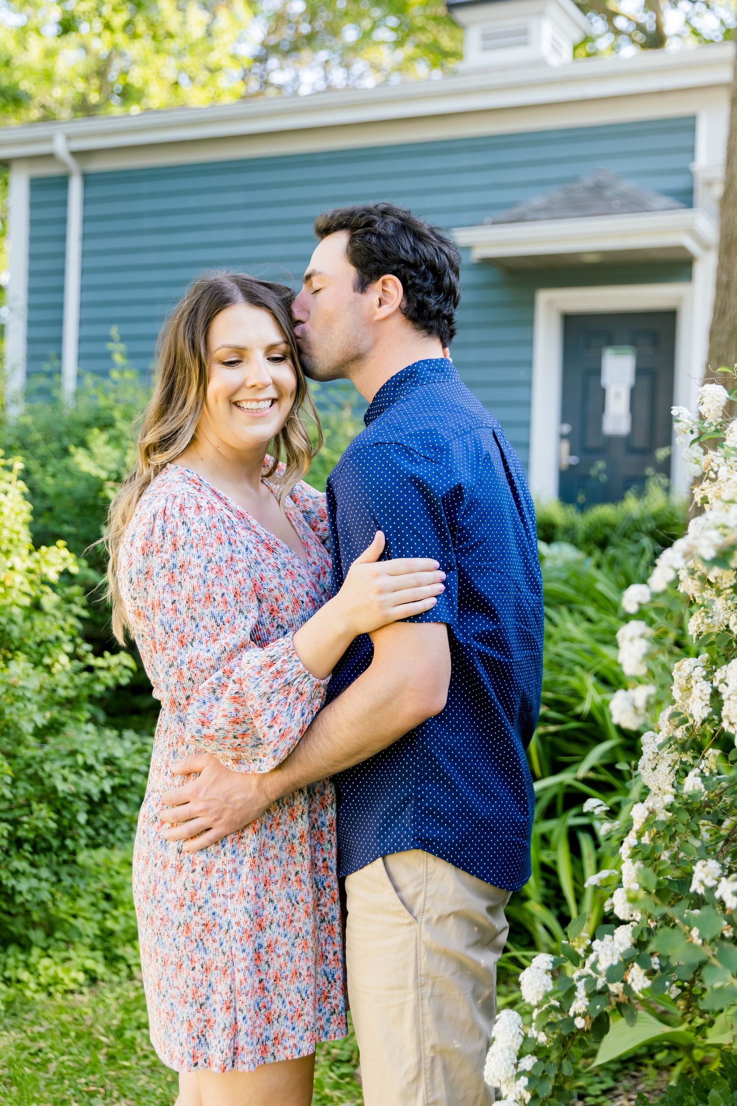 Intimate moment between couple in front of blue house surrounded by flowering bushes.