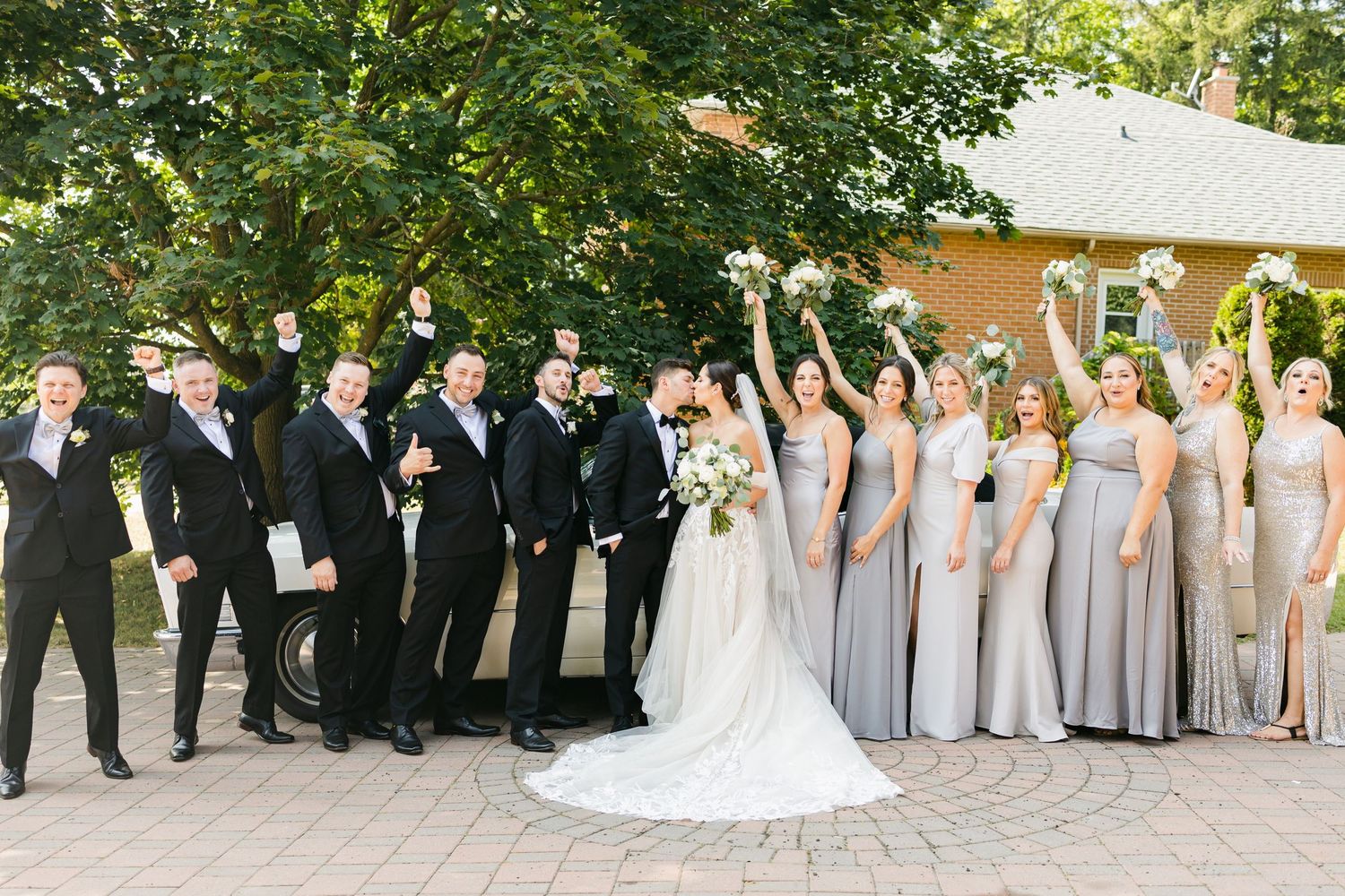 Joyful wedding party celebration shot with bridesmaids in silver and groomsmen in black tuxedos.