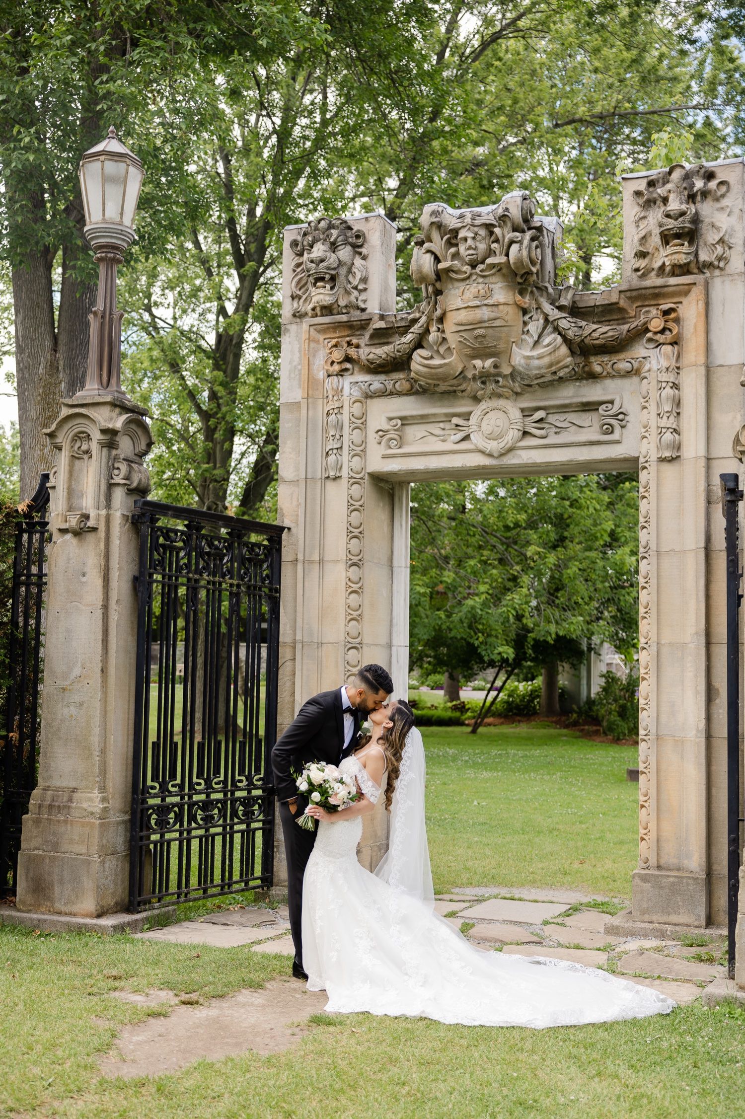 Wedding couple shares an intimate moment under a decorative stone gateway with iron gates and lanterns.