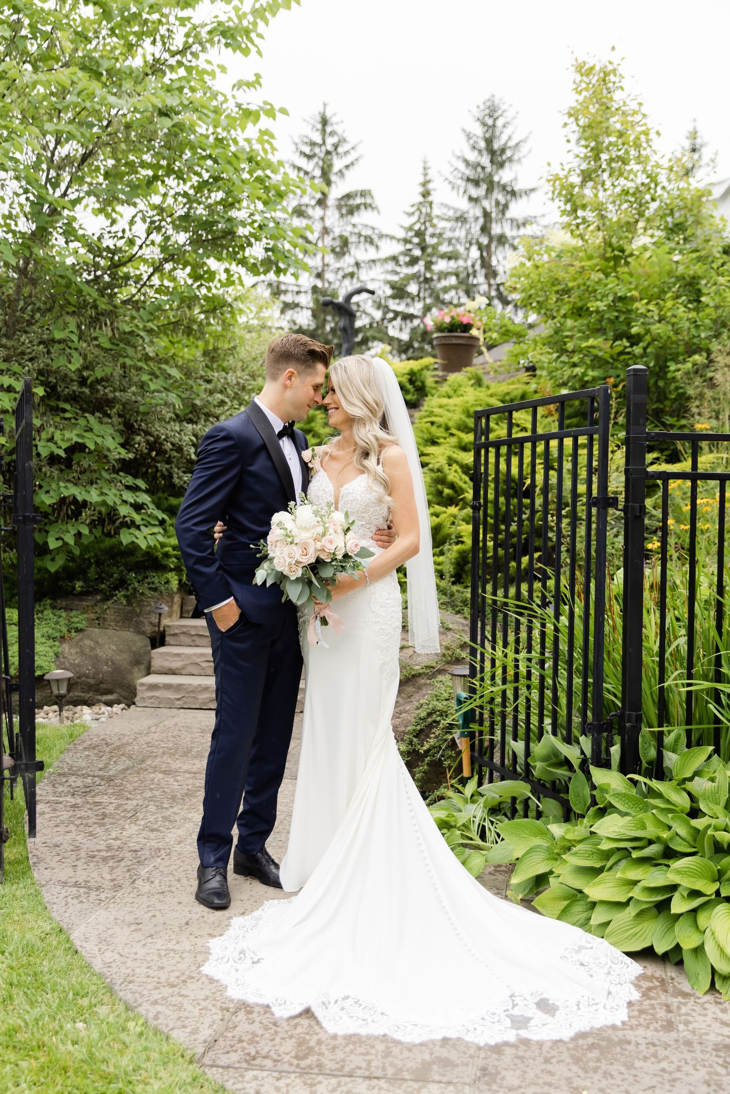 Couple embraces on stone garden path with elegant wedding attire and floral bouquet.