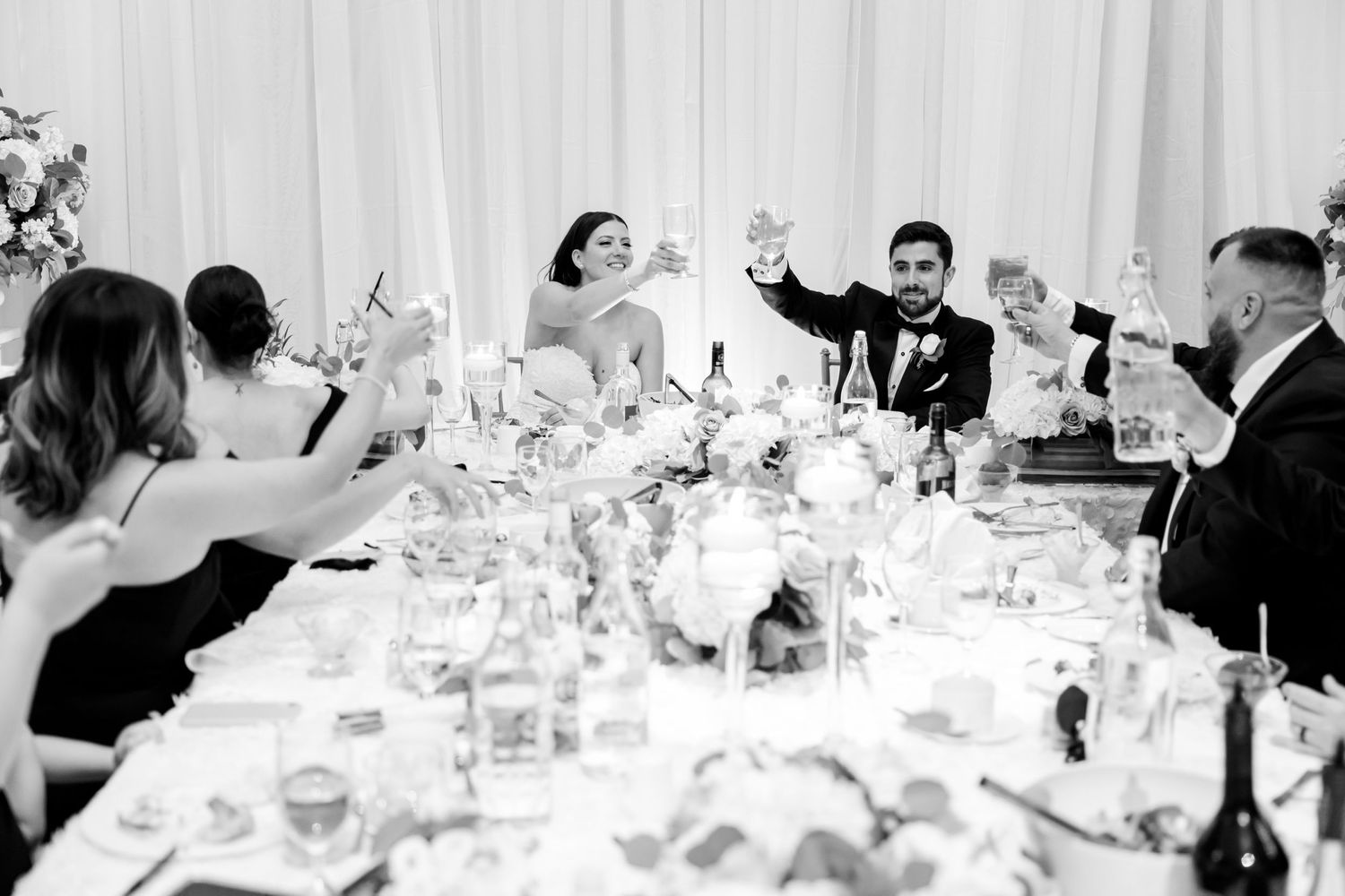 Black and white photo of wedding reception toast with guests raising glasses.