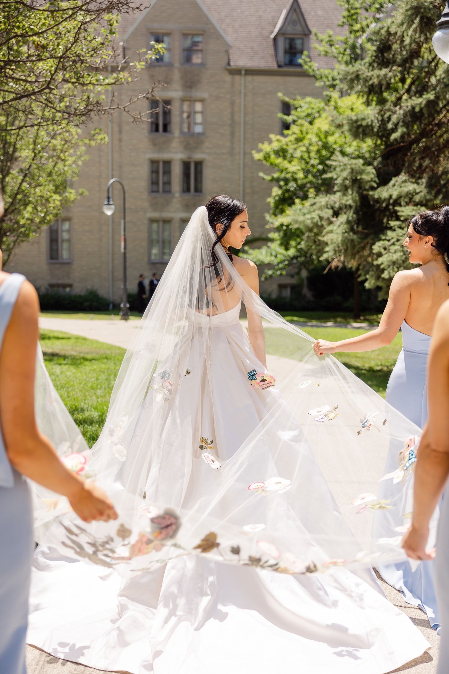 Bridesmaids in light blue dresses help arrange bride's flowing veil outdoors.