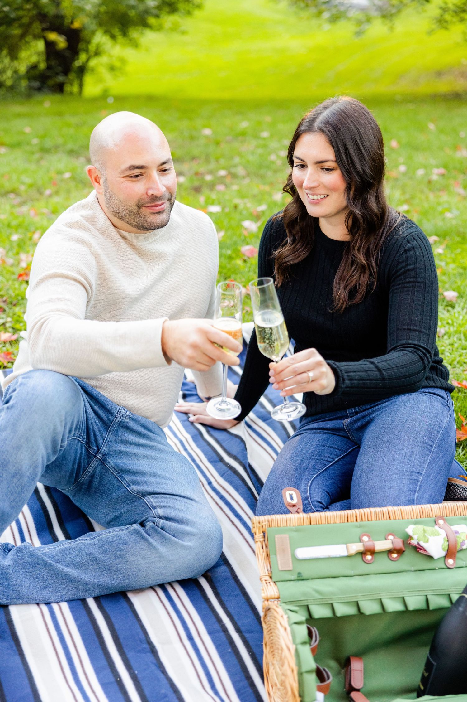 Couple toasts with champagne while having a romantic picnic on a striped blanket in a lush green park.