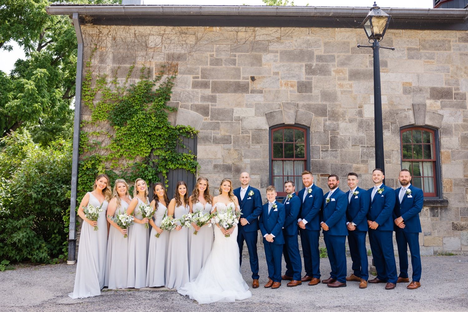 Large wedding party in navy and white attire poses against historic stone building.
