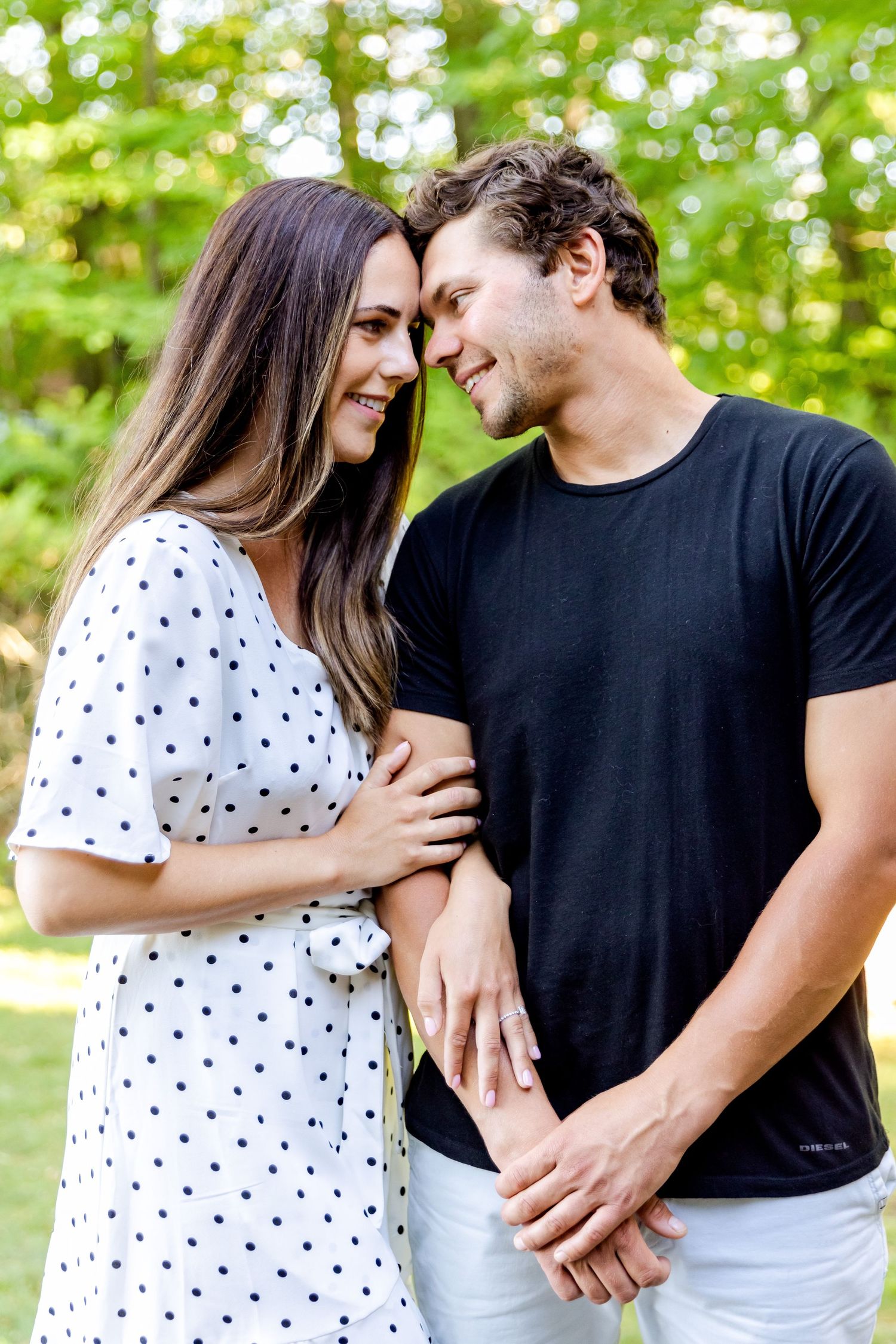 A couple shares an intimate moment outdoors wearing a polka dot dress and black t-shirt against green foliage.