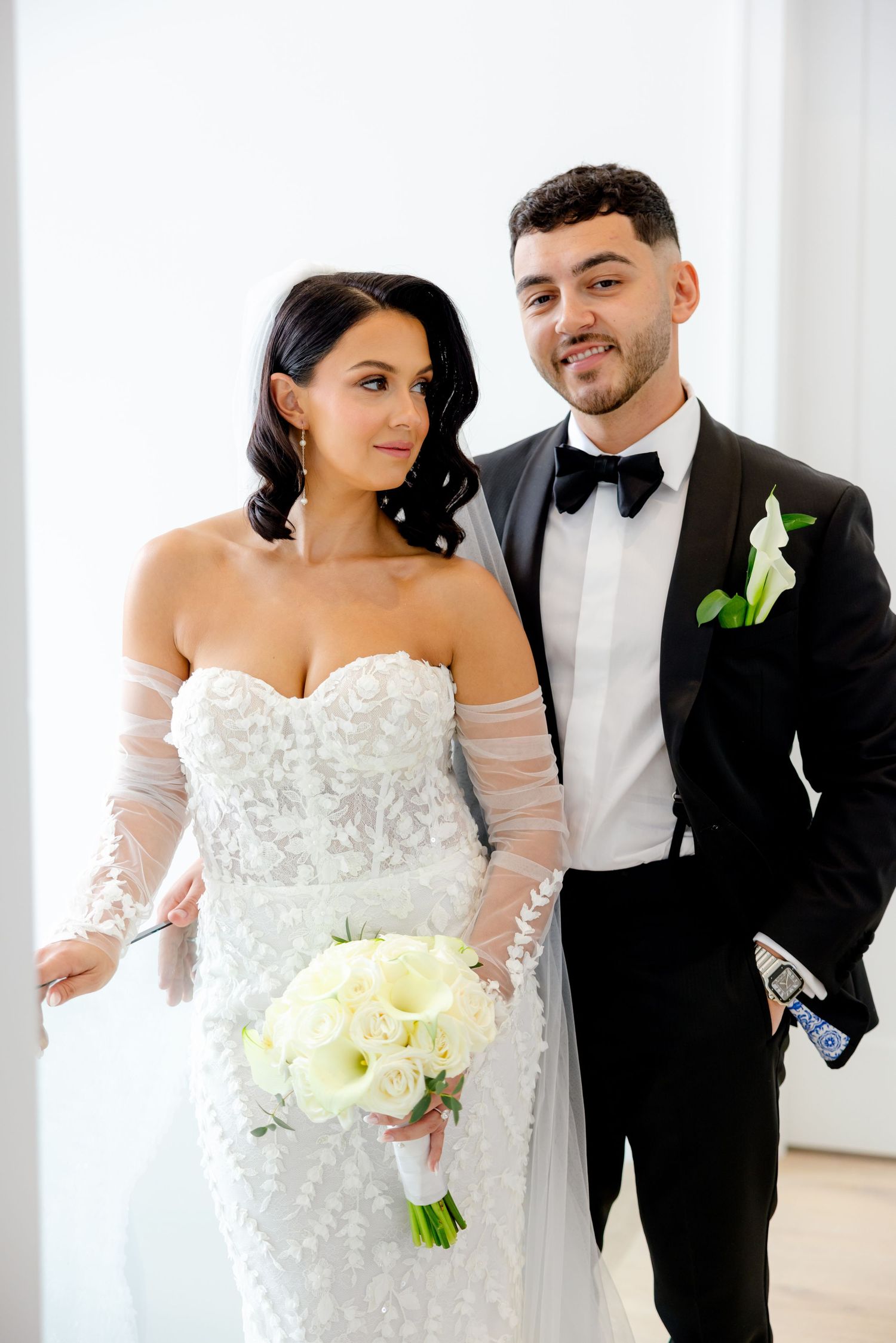 Wedding couple in classic attire poses against white interior with yellow bouquet.