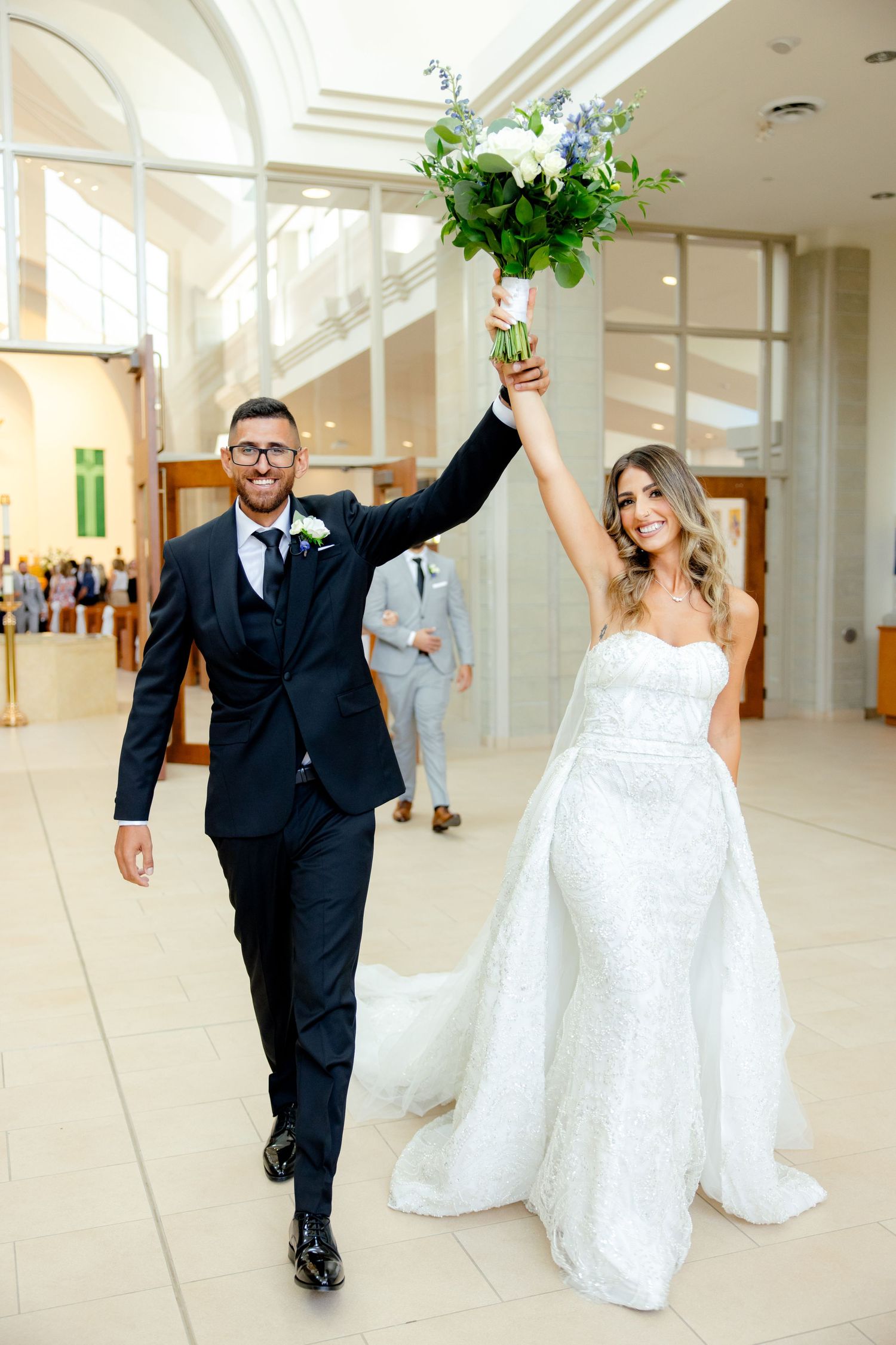 Joyful couple celebrates in church aisle as groom holds up white flower bouquet triumphantly.