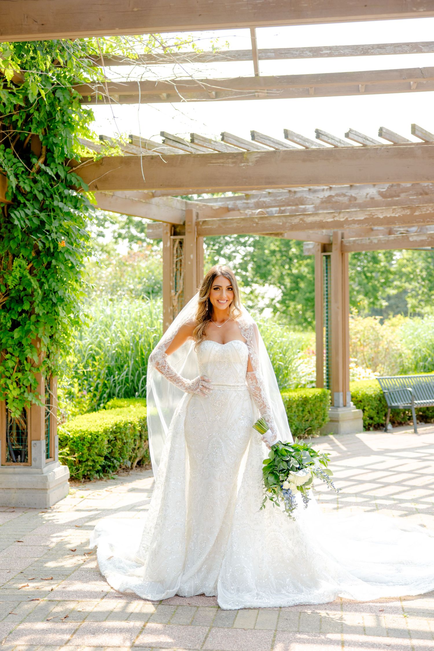 Sun-drenched bridal portrait showcasing fitted lace wedding dress and cathedral veil under pergola.
