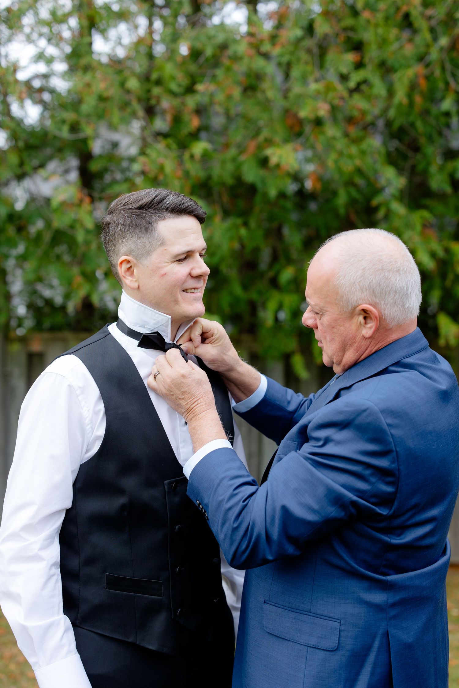 Two people share a candid moment while adjusting formal attire outdoors.
