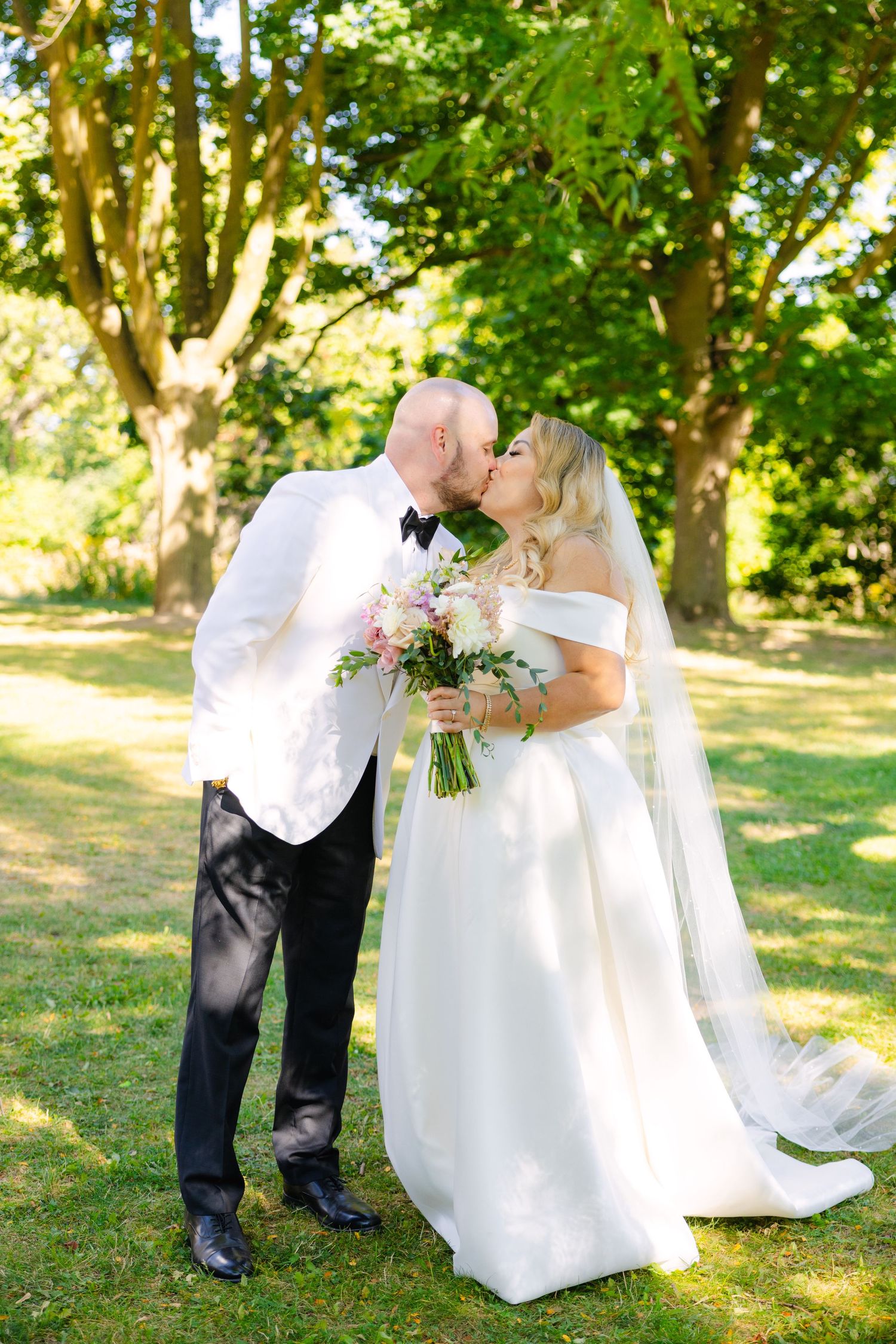 Romantic wedding portrait of couple sharing a kiss outdoors with bride holding floral bouquet.