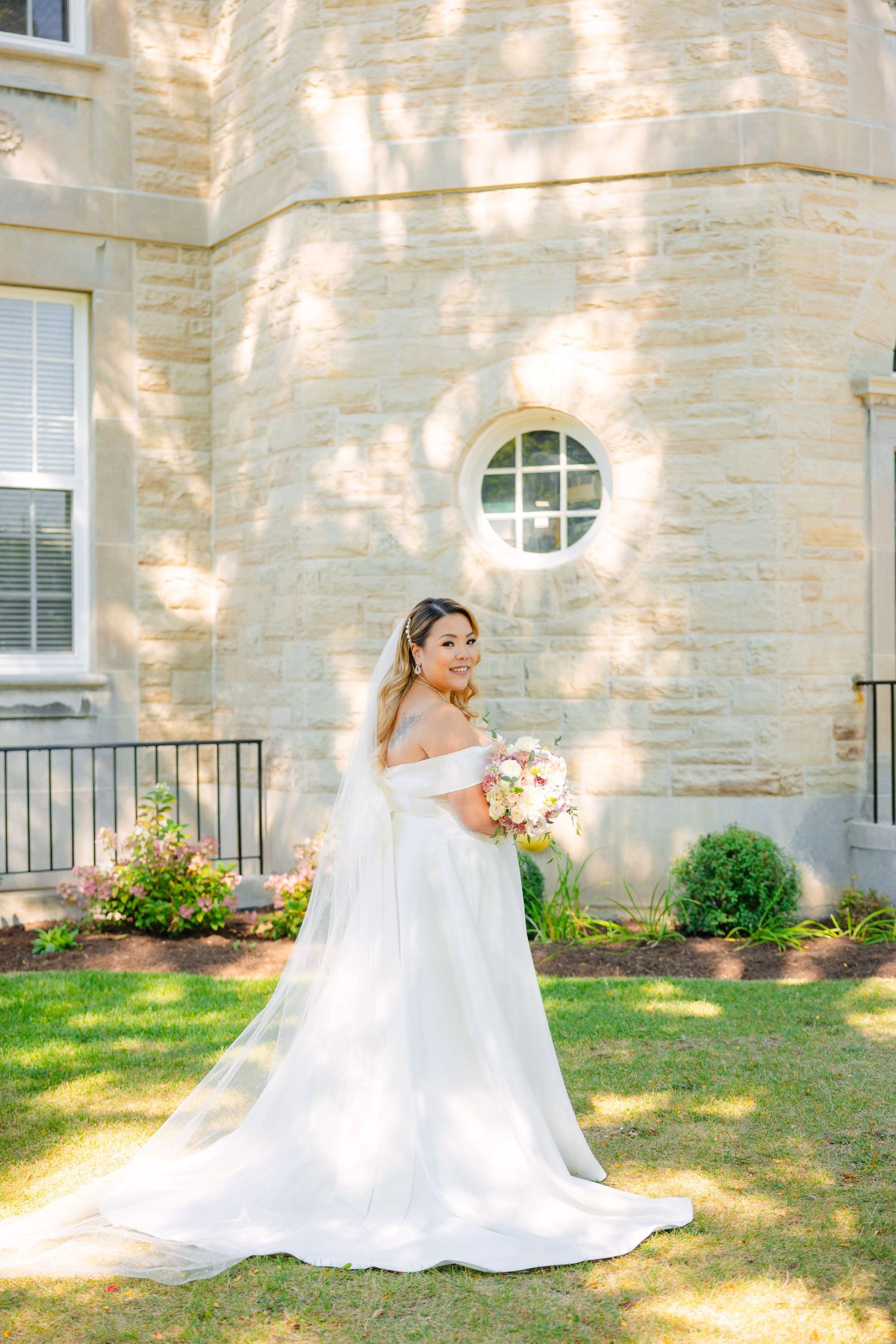 Beautiful bride in flowing white gown poses against stone building with round window on sunny day.