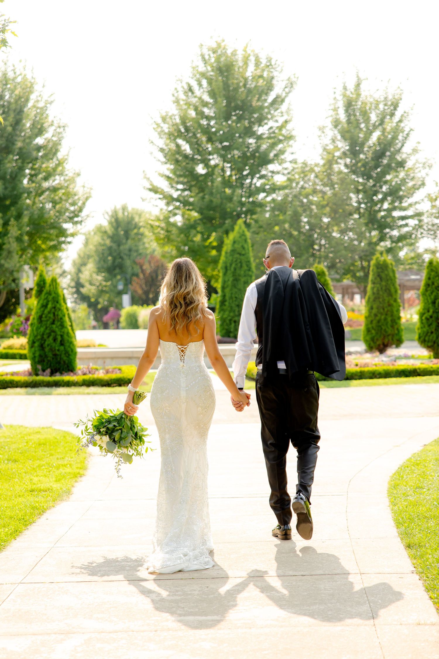 A bride in a fitted white gown and groom in black suit walk hand-in-hand down a garden path lined with manicured trees.