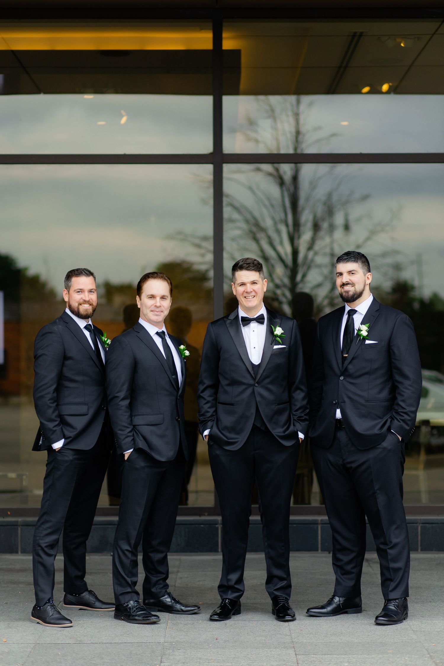 Groomsmen in dark suits pose together outside modern building with reflective windows at dusk.