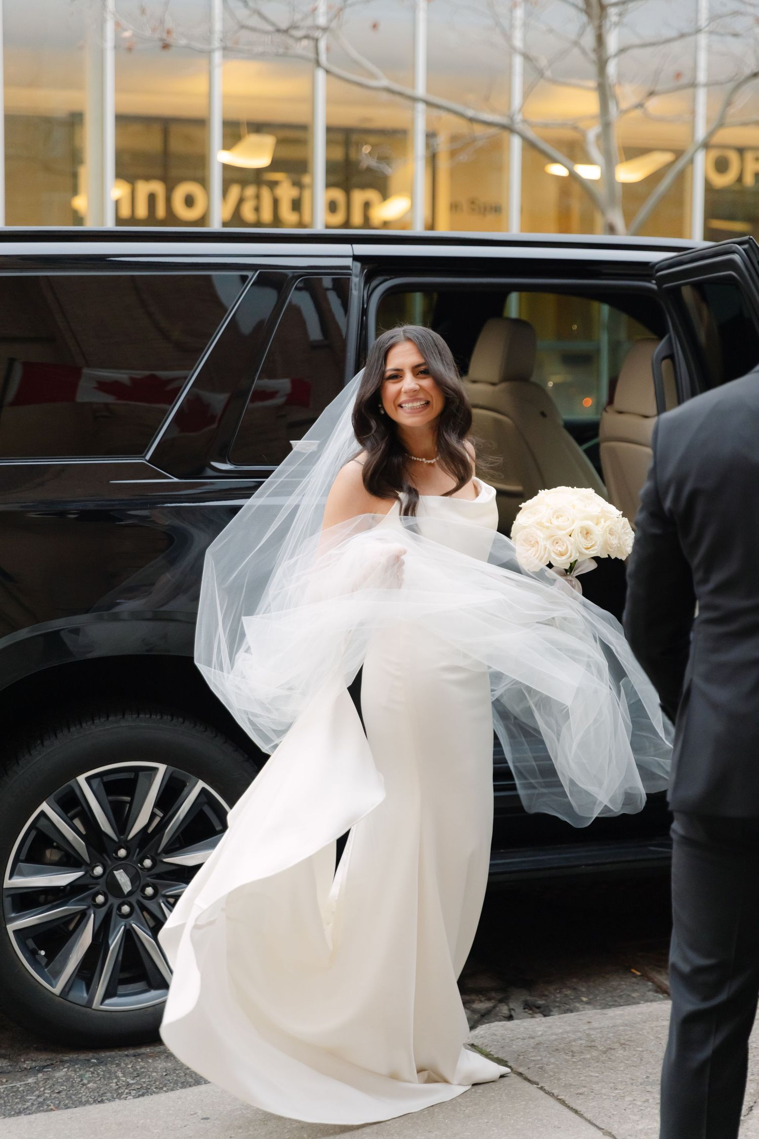Elegant bride in flowing white dress stepping out of luxury black car.