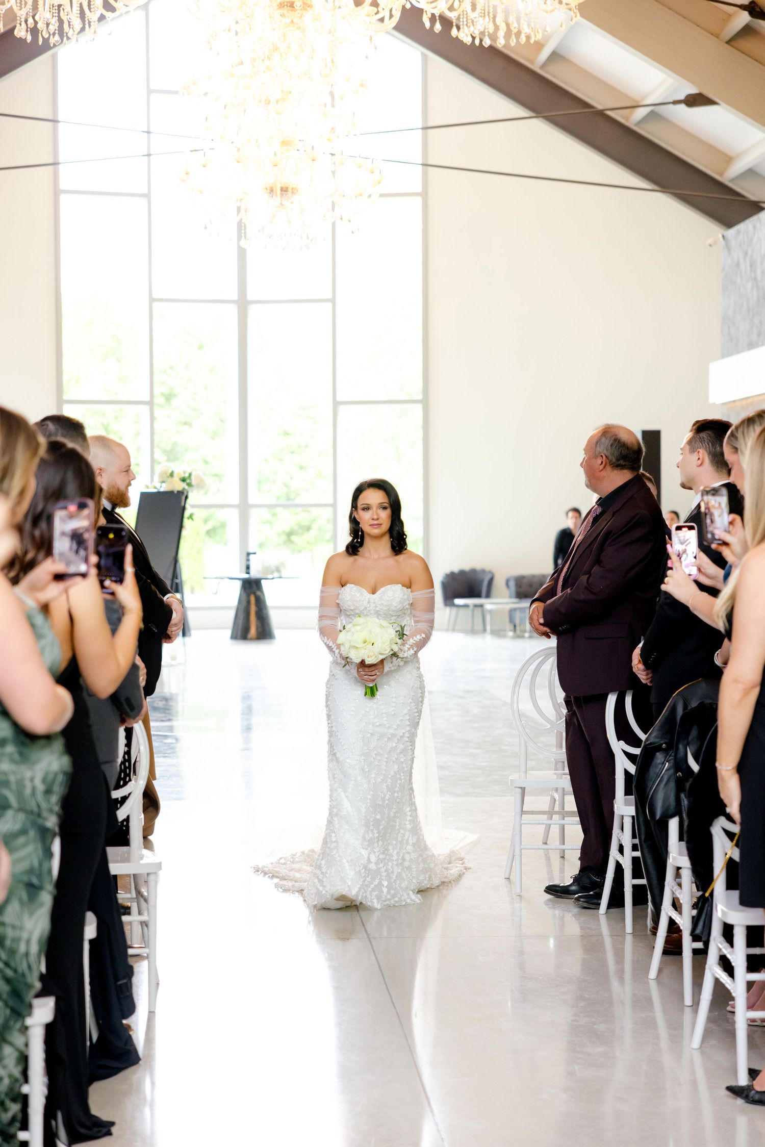 Bride in white gown walks down aisle in modern church with guests watching from sides.