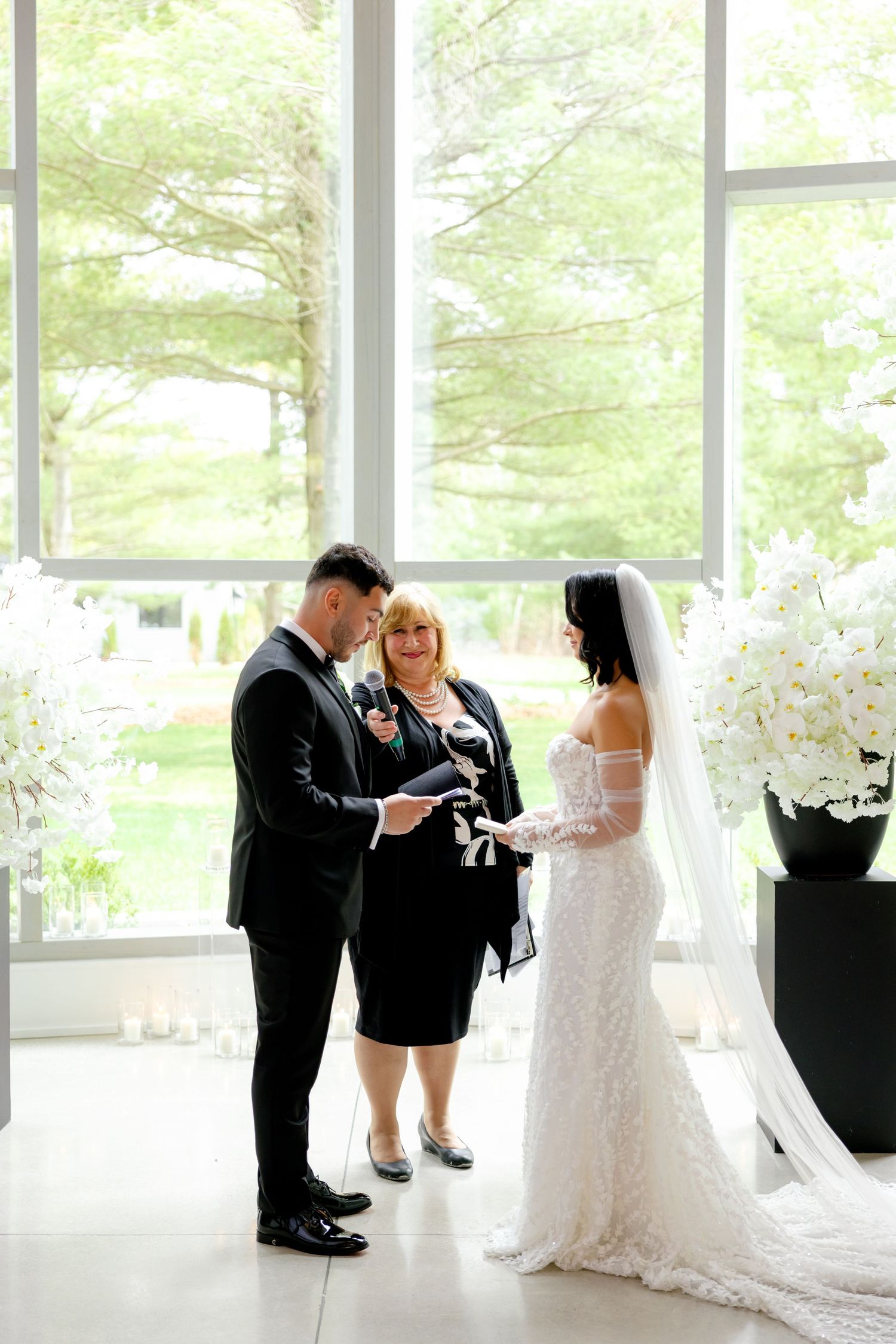 Wedding ceremony taking place in bright sunlit room with tall windows and white floral arrangements.
