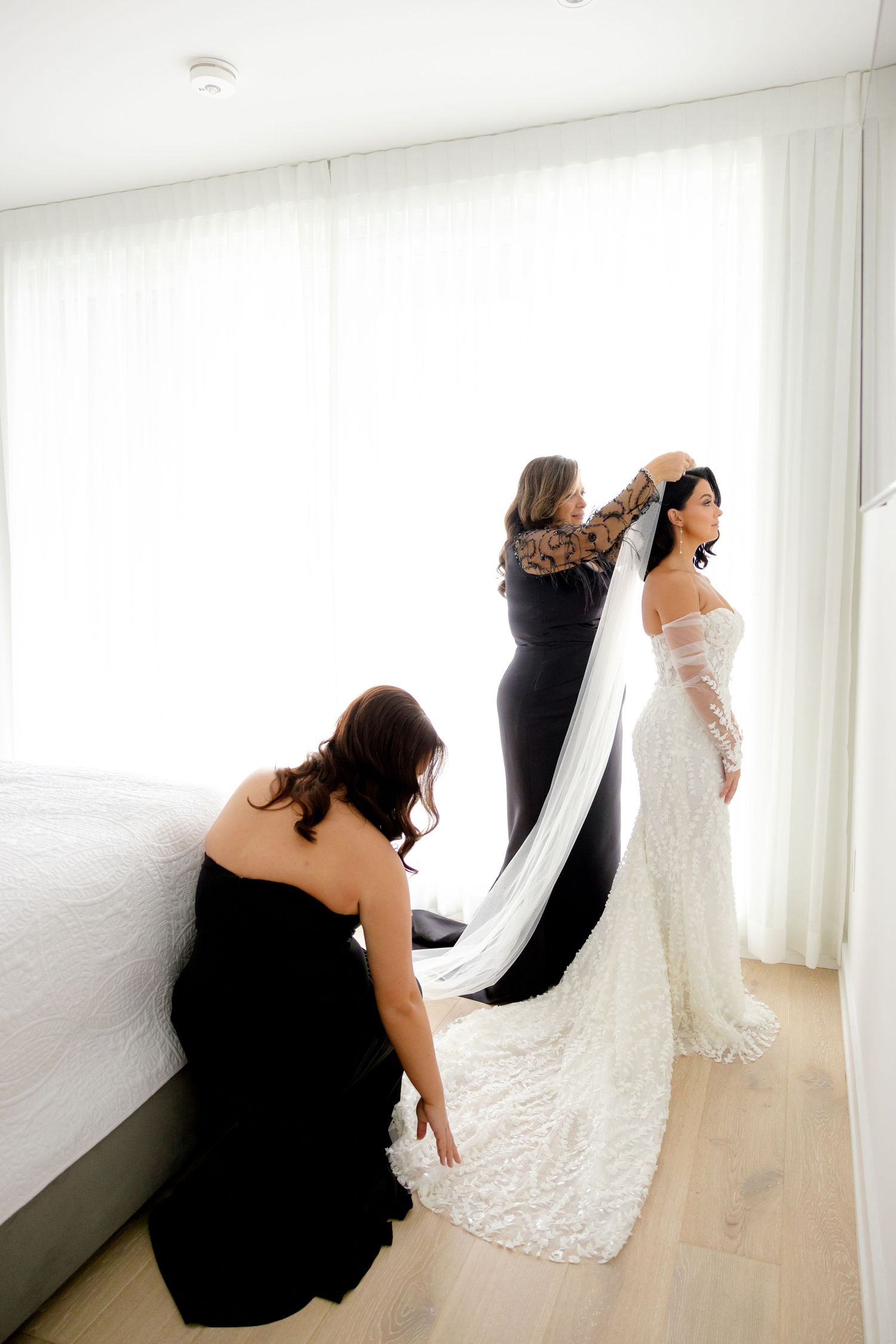 Two women help adjust the bride's long veil in a bright white room during wedding preparations.