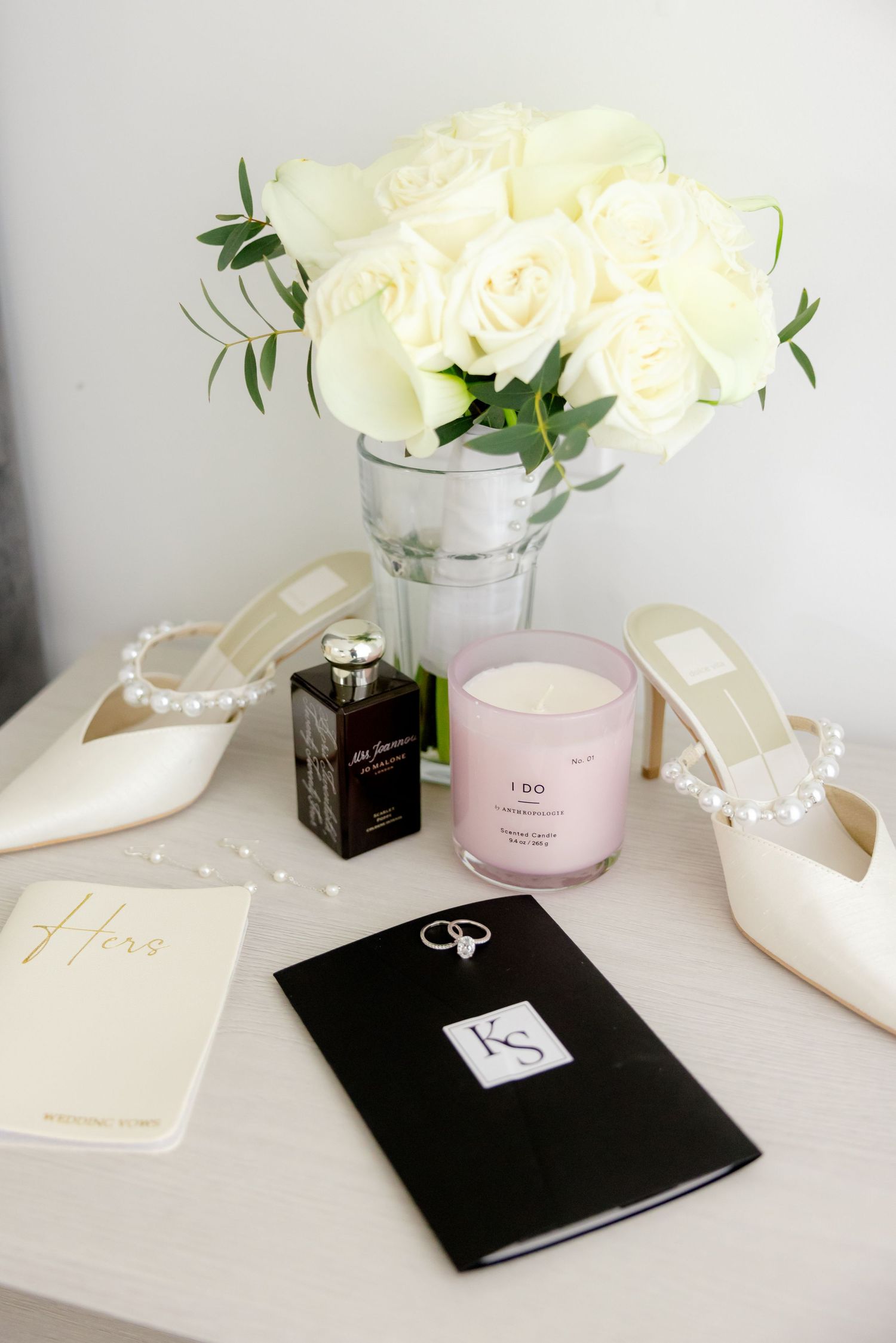 White roses in glass vase with wedding accessories including perfume, candle and shoes on white surface.