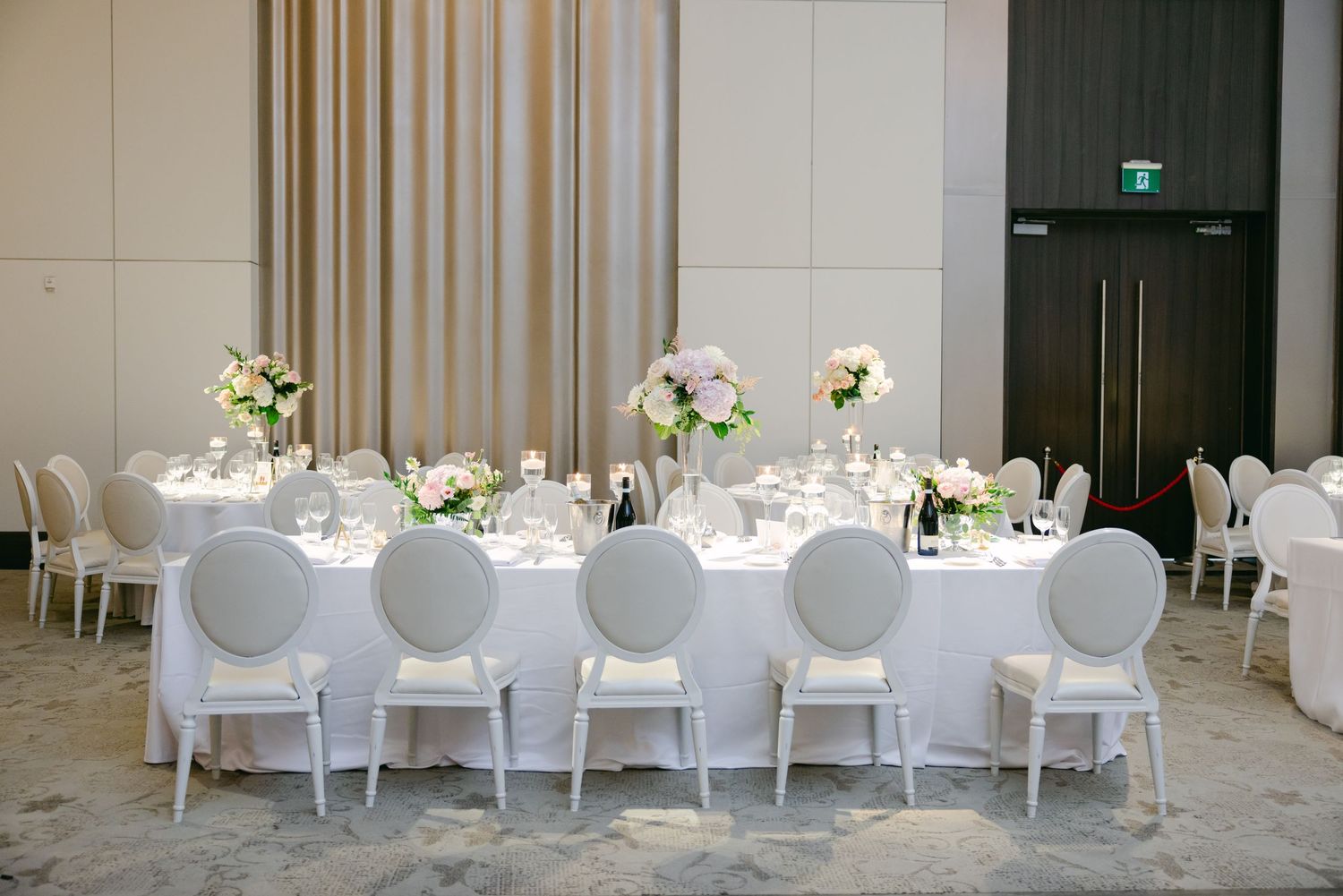 Long wedding reception table with white oval-back chairs and classic floral centerpieces.