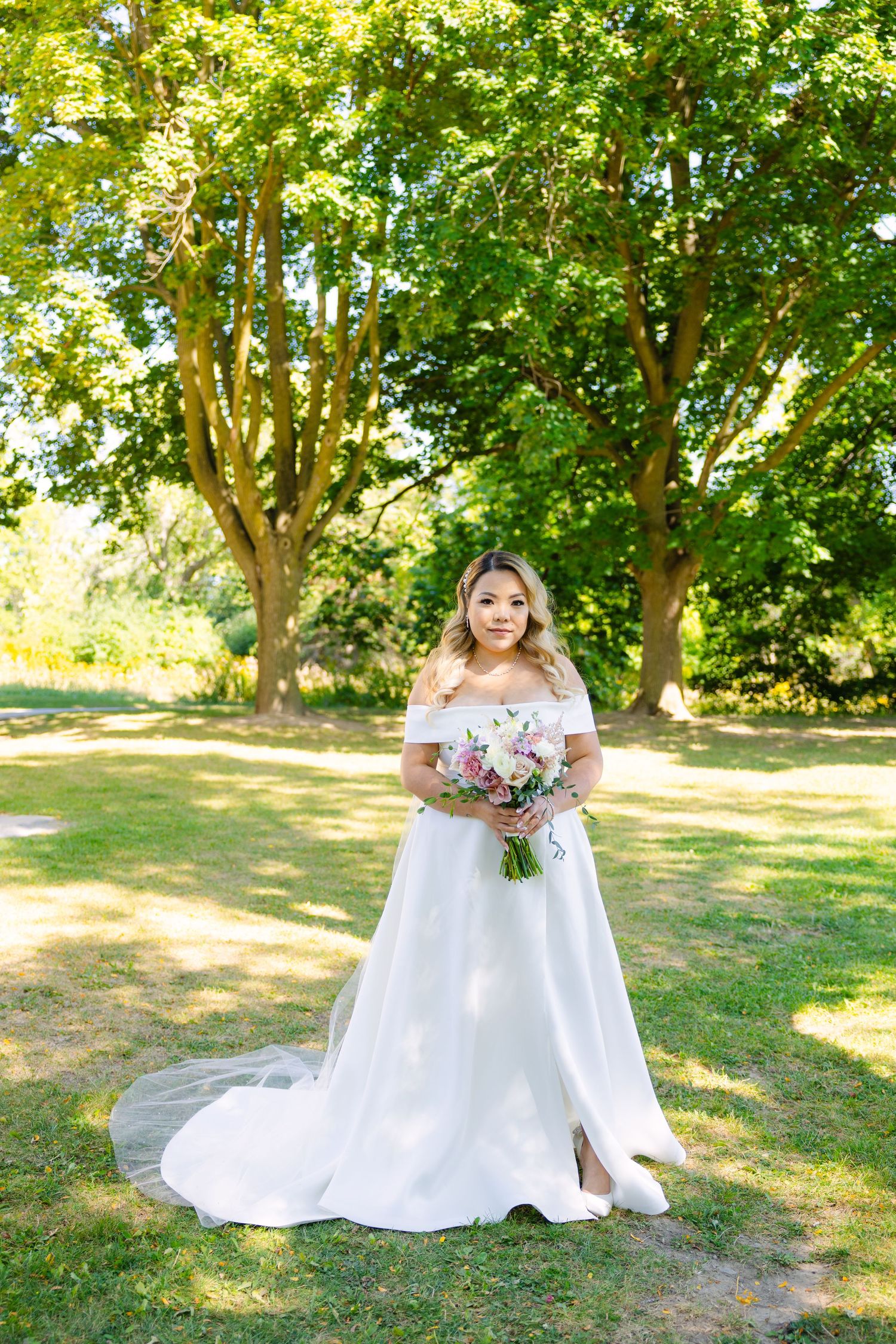 Full-length portrait of bride in flowing white gown holding bouquet in garden setting.