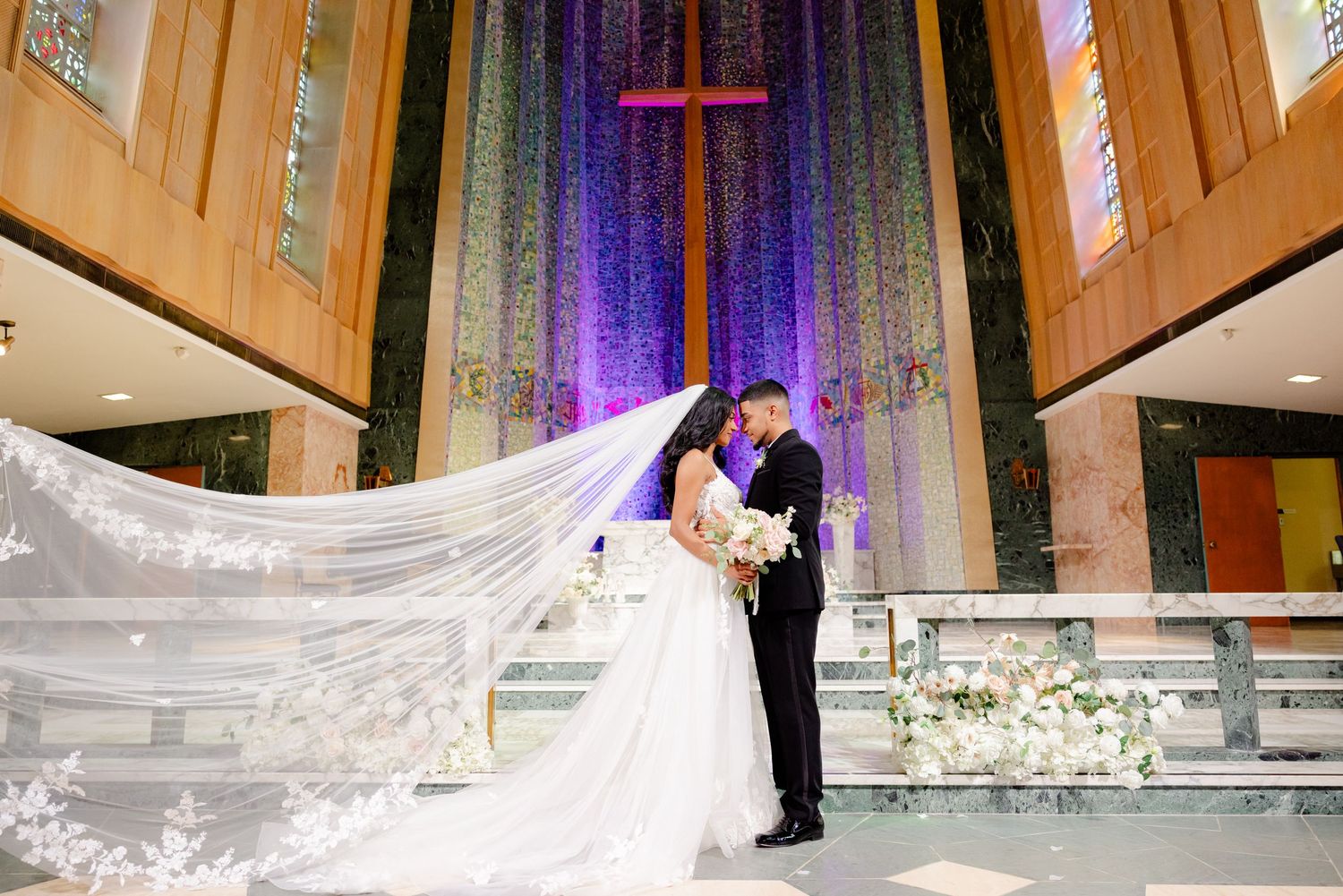 Elegant church wedding photo with dramatic veil sweep and purple cross backdrop.