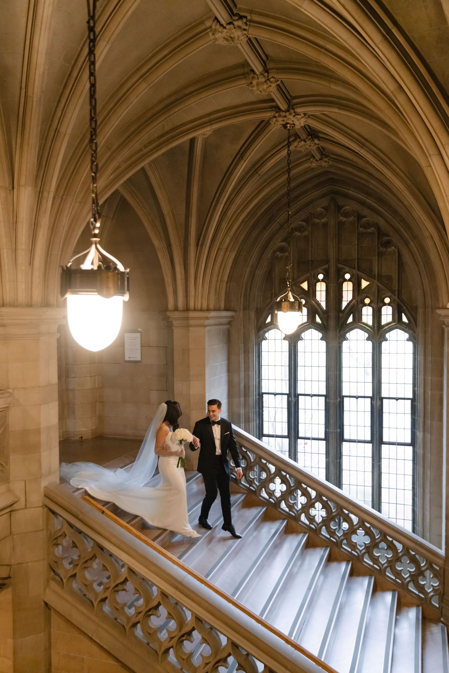 Wedding couple poses on grand stone staircase under gothic arches and hanging pendant light.
