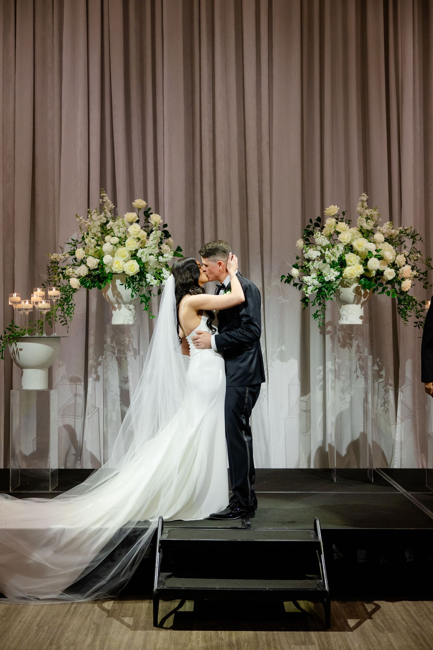 Couple embracing on stage with elegant floral arrangements and draped backdrop at wedding ceremony.